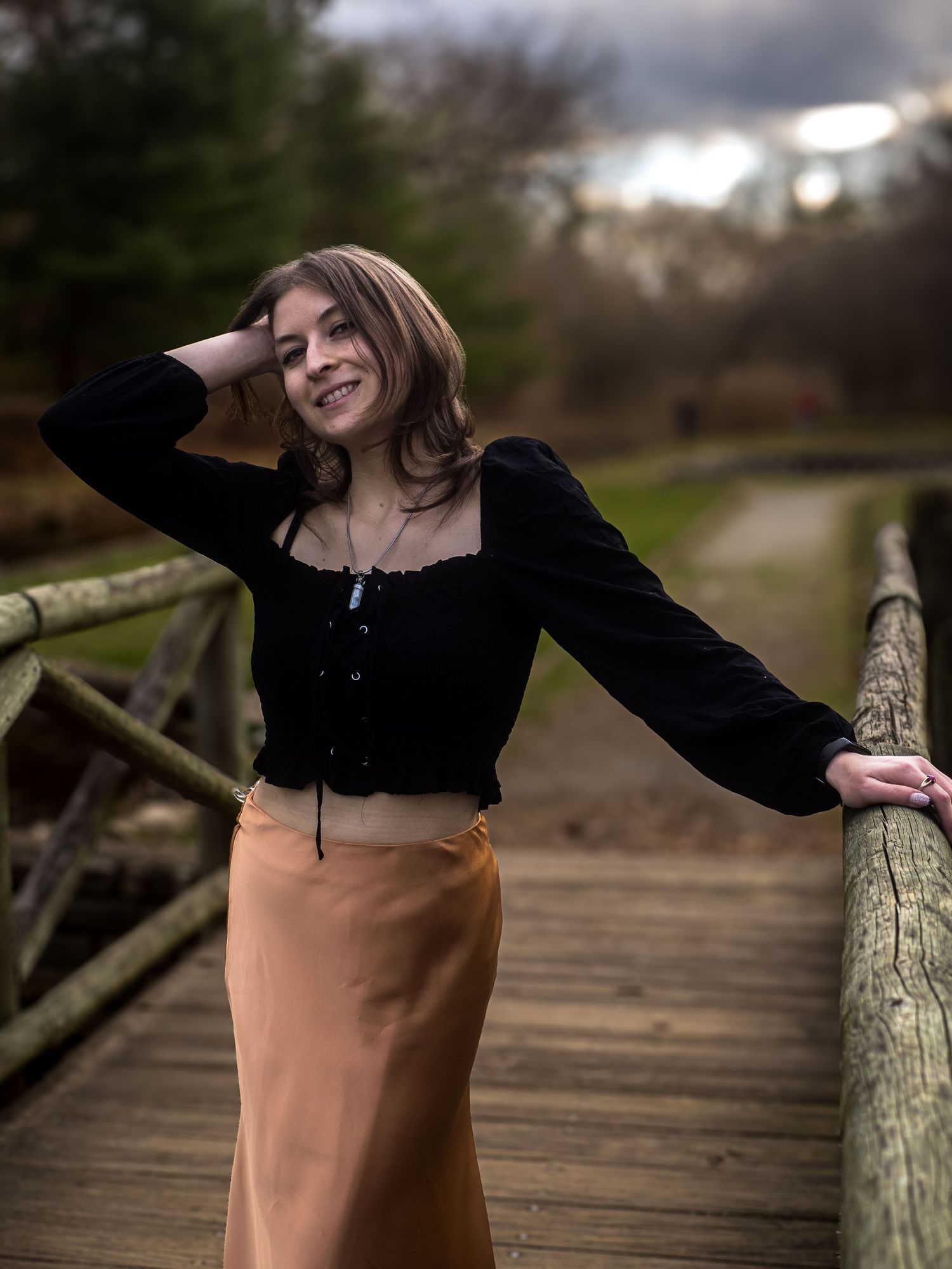 Person in black long sleeve top and peach skirt poses on wooden bridge in natural outdoor setting.