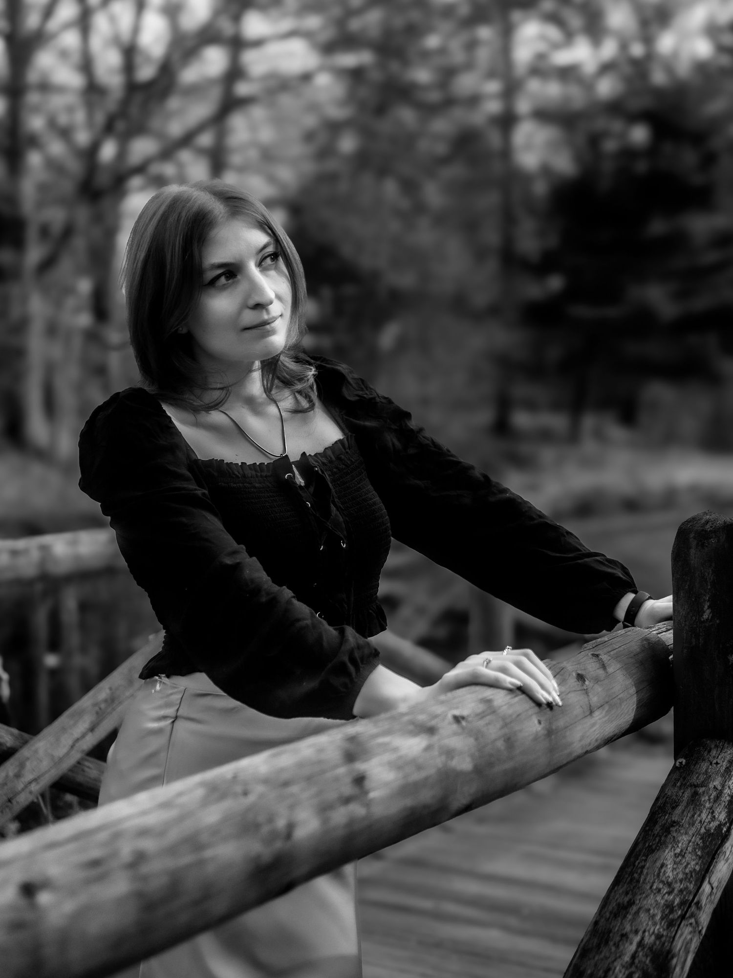 A black and white portrait shows a person in vintage attire leaning on a wooden railing in a forest setting.