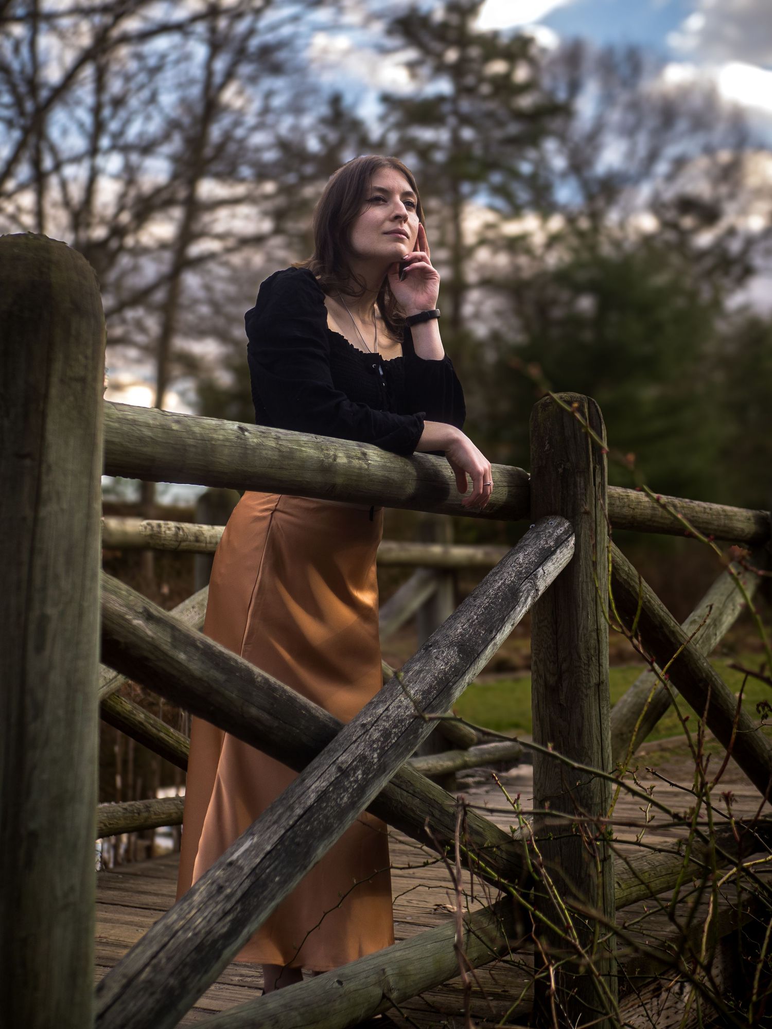 A person in a black top and brown skirt leans thoughtfully against a rustic wooden fence in a natural outdoor setting.