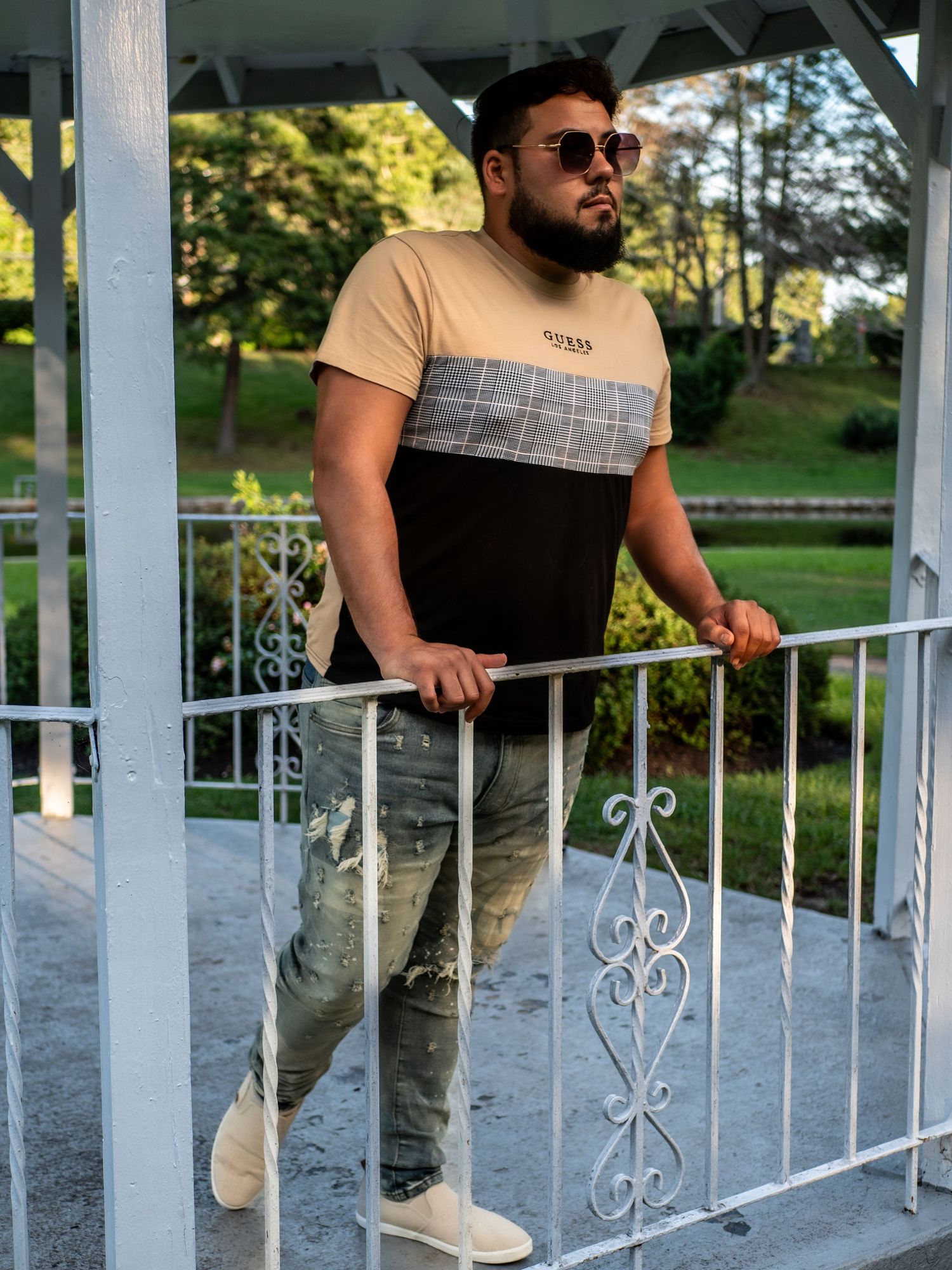 A casually dressed person leans against a white ornate porch railing on a sunny day wearing sunglasses.