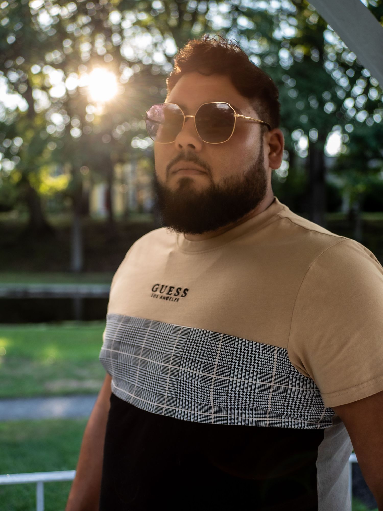 A person wearing sunglasses and a tan, plaid and black color-blocked t-shirt stands outdoors in natural sunlight.