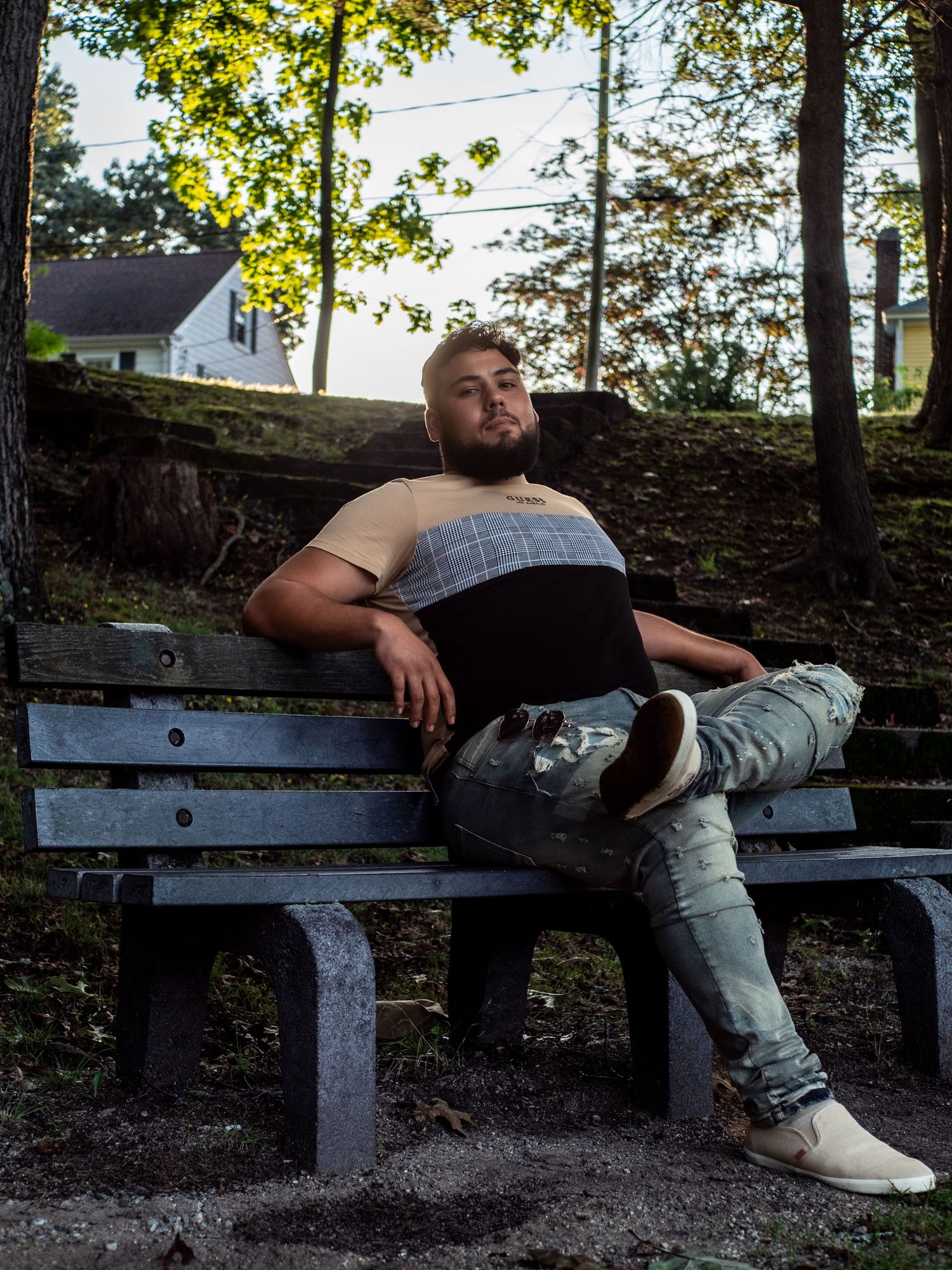A person relaxes on a park bench surrounded by trees and greenery, wearing casual clothing and sneakers.