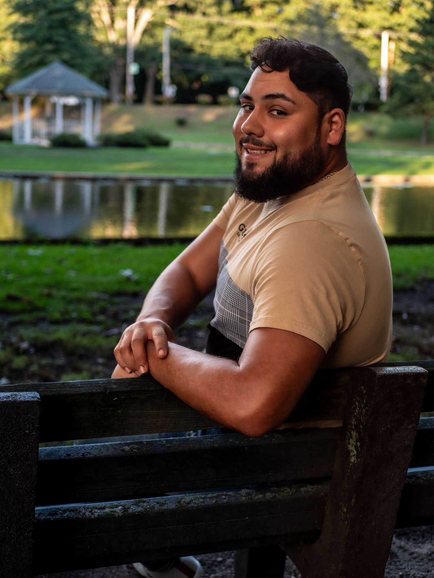 A person relaxes on a wooden bench by a scenic pond with a gazebo visible in the background during golden hour.