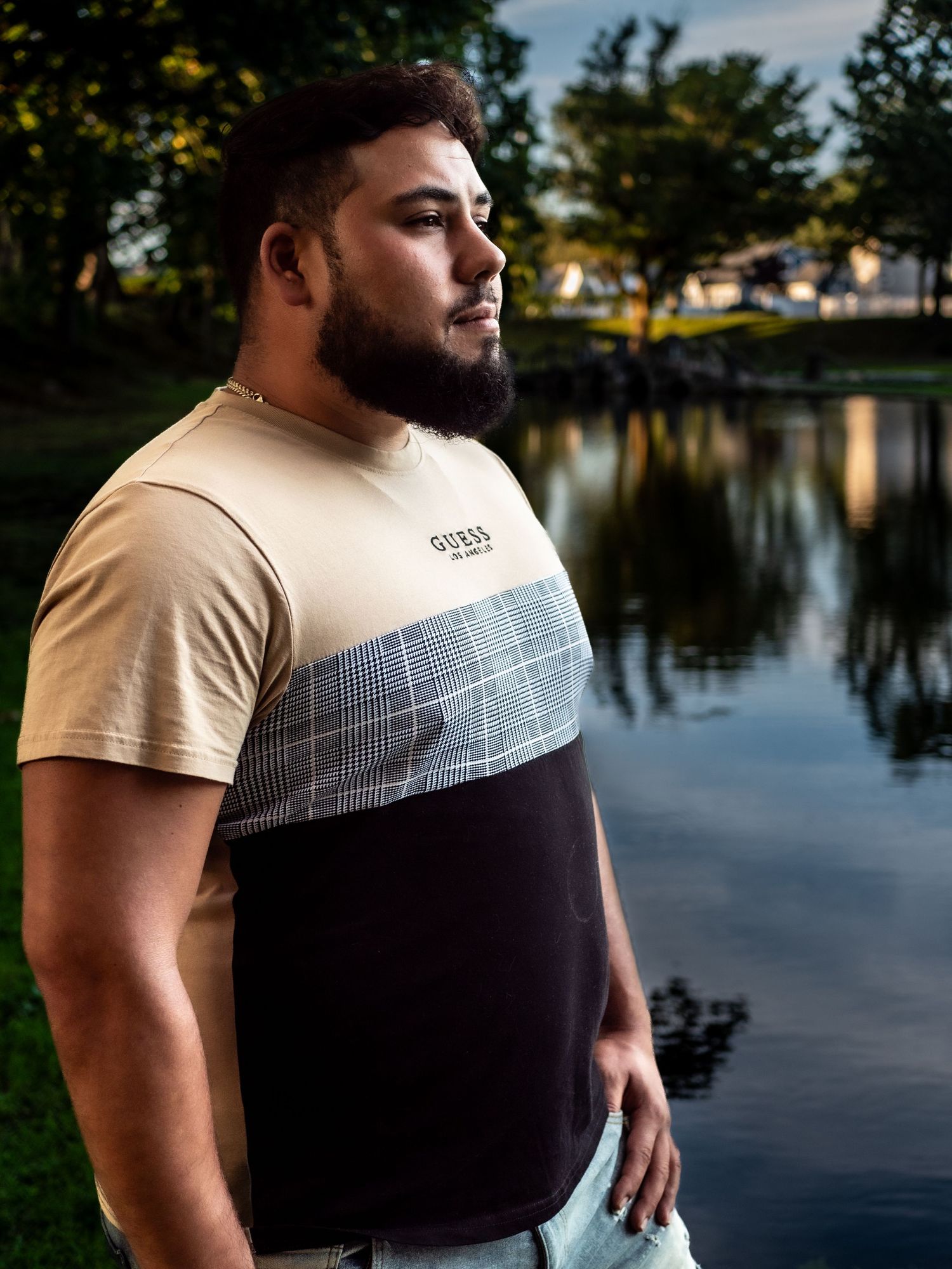 Person wearing a color-block t-shirt stands near a tranquil pond at sunset with greenery in the background.