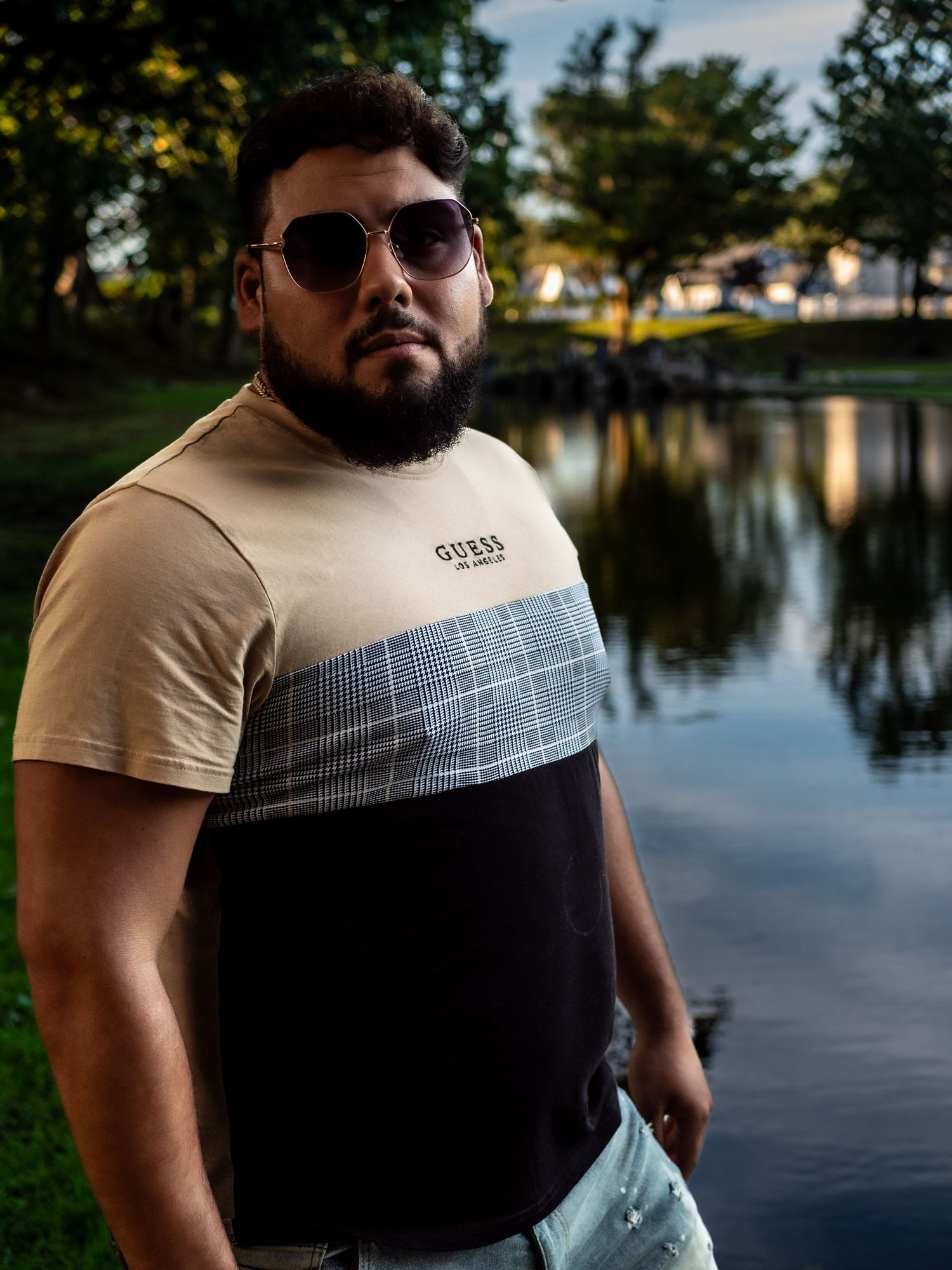 Portrait of person wearing sunglasses and two-tone t-shirt standing by a serene lake during golden hour.