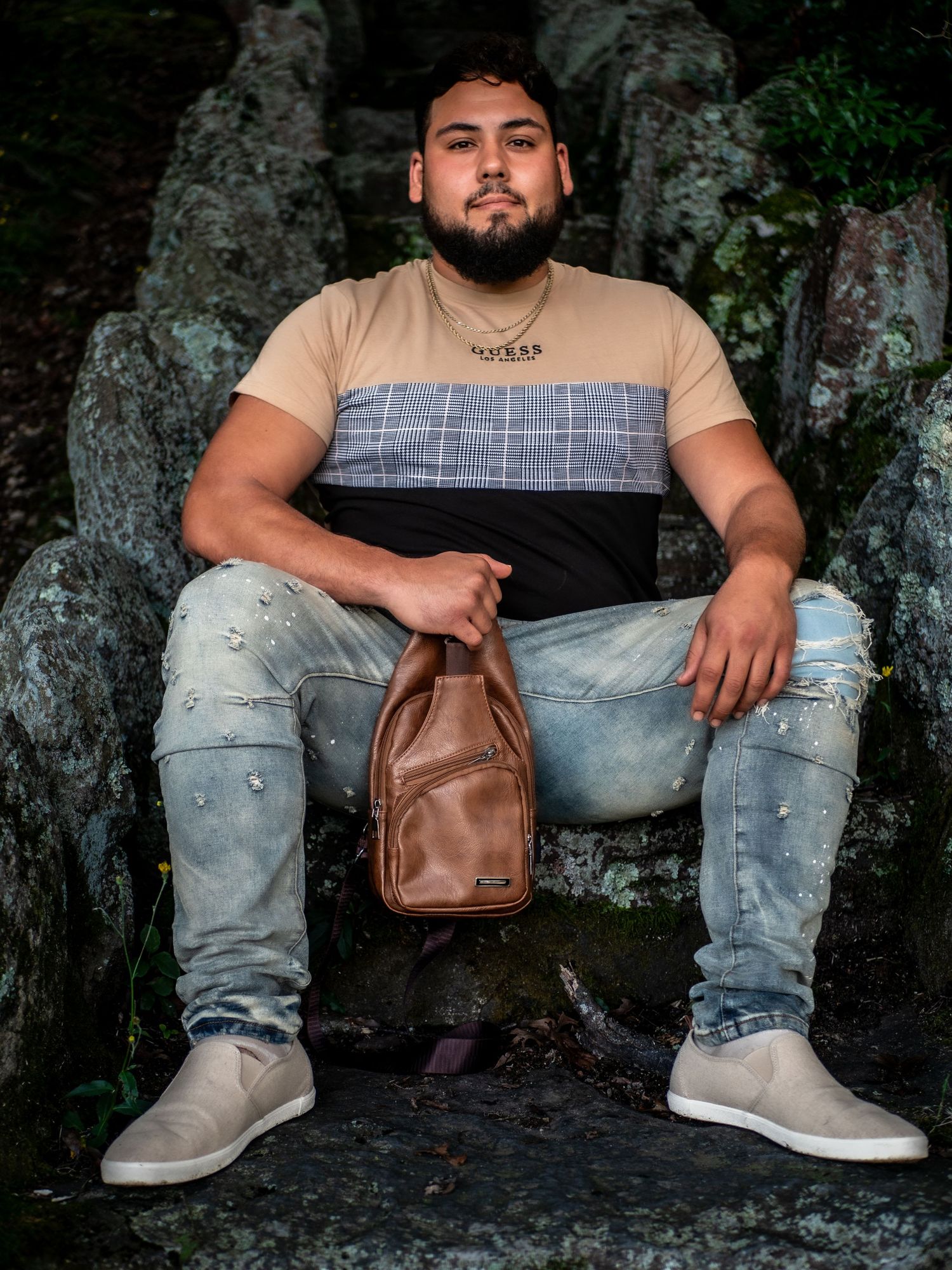 Man in beige and black color block shirt sits on rocks with distressed jeans and tan shoes, holding brown leather bag.