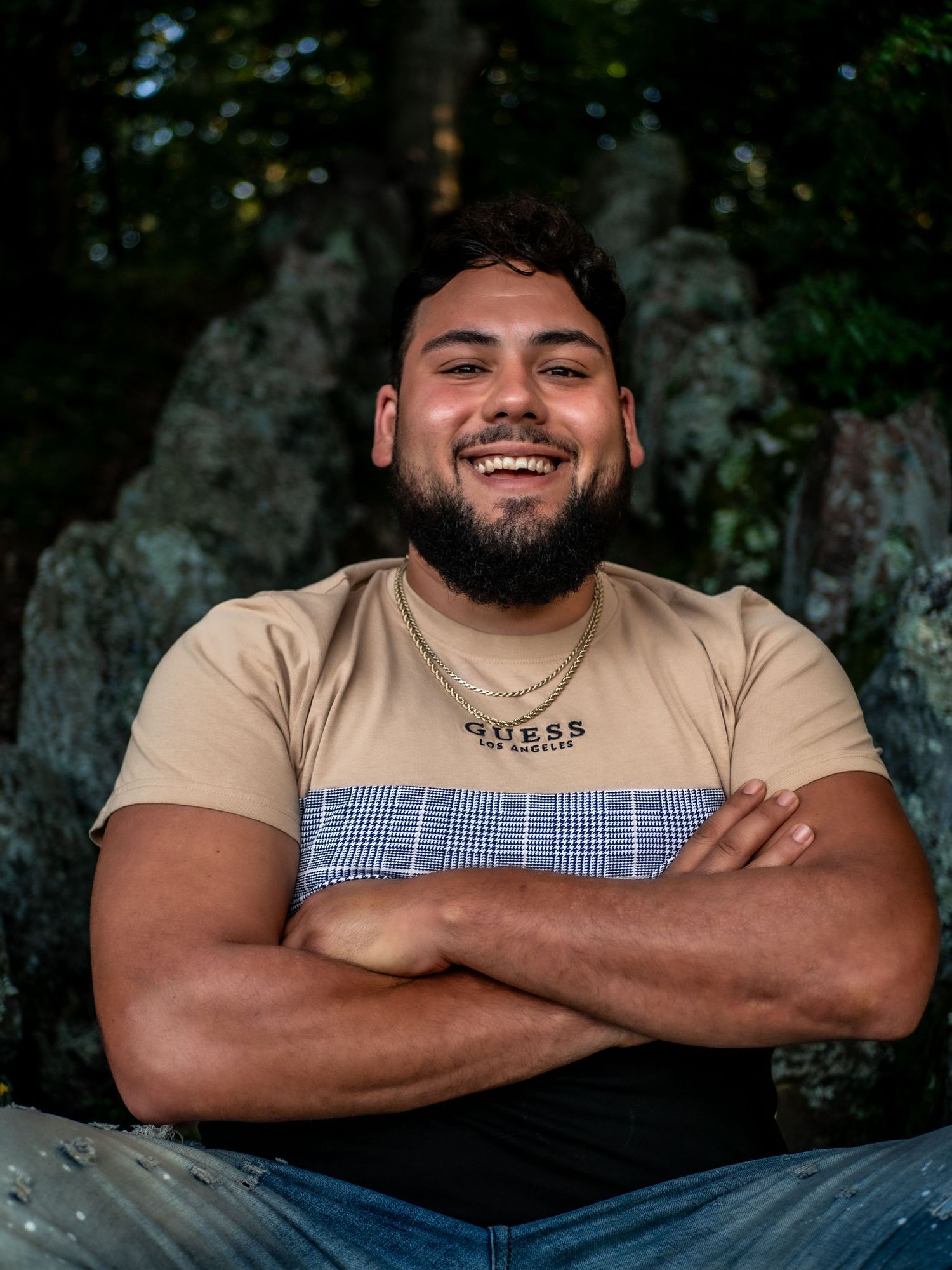 A joyful portrait of someone wearing a tan and blue color block t-shirt with arms crossed against a dark background.