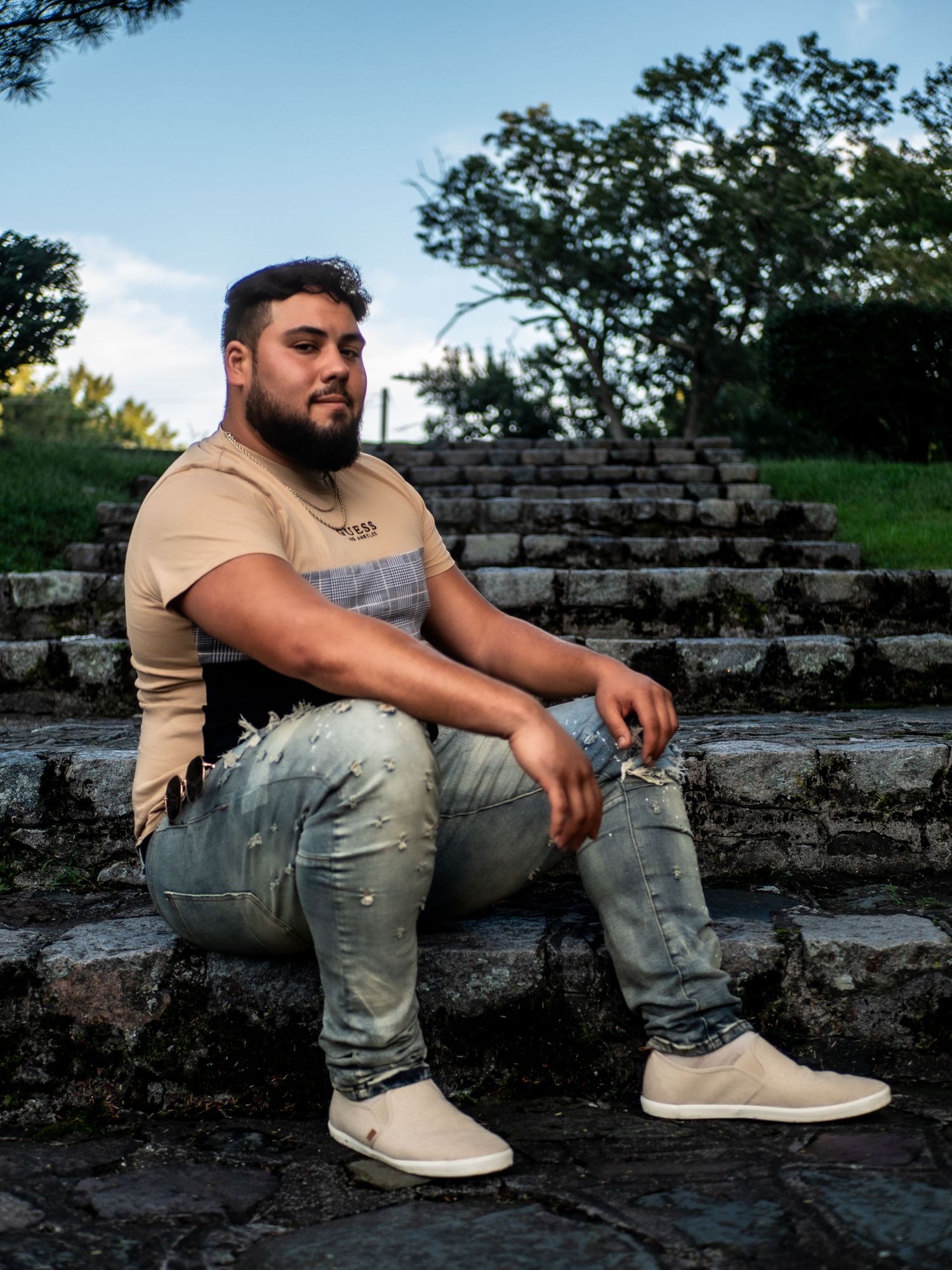 A person sits thoughtfully on stone steps in a park wearing a beige t-shirt, distressed jeans and light colored sneakers.