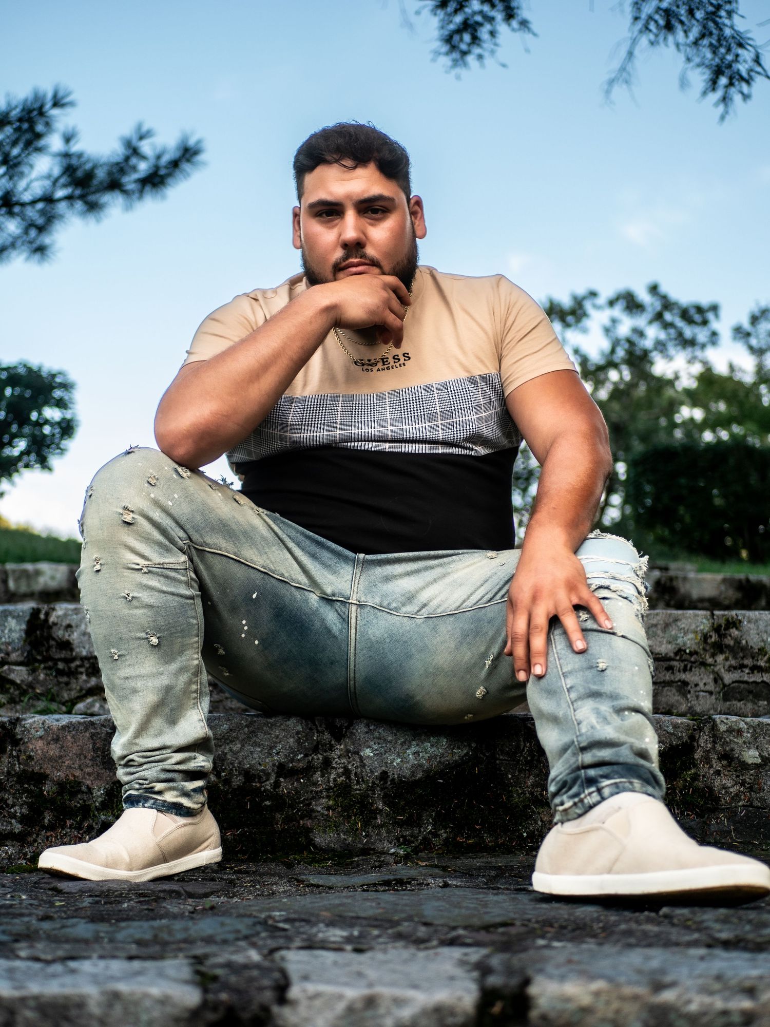A candid portrait shot of someone in a striped shirt and distressed jeans sitting on stone steps outdoors.