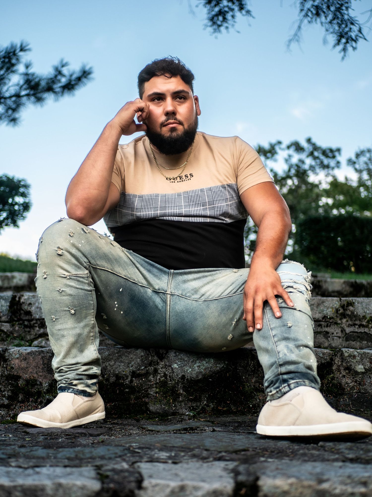 Person wearing a tan, grey and black colorblock t-shirt and light jeans sits on concrete steps in an outdoor setting.