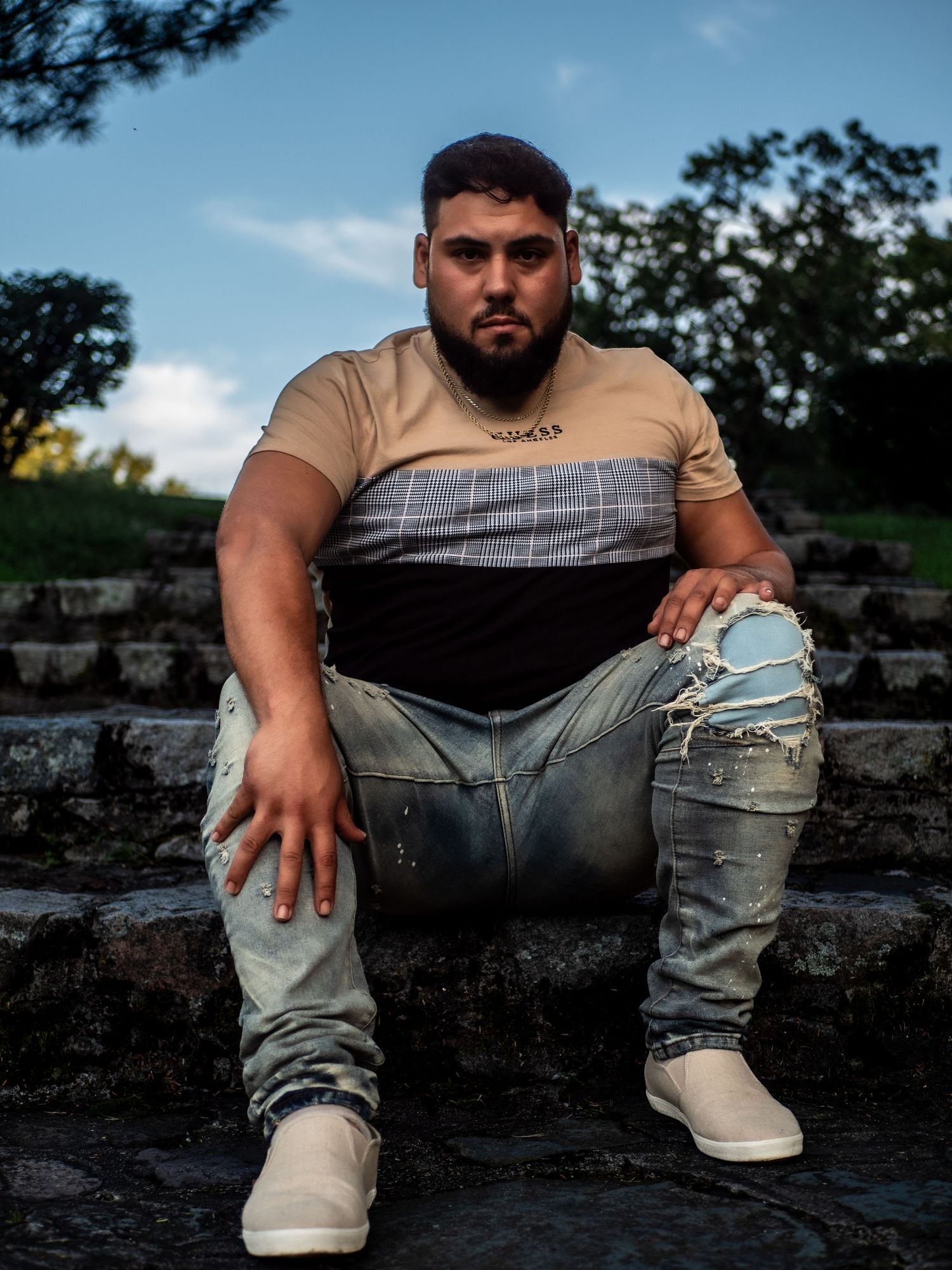 A person in a beige and black striped shirt and distressed jeans sits on stone steps in an outdoor setting.