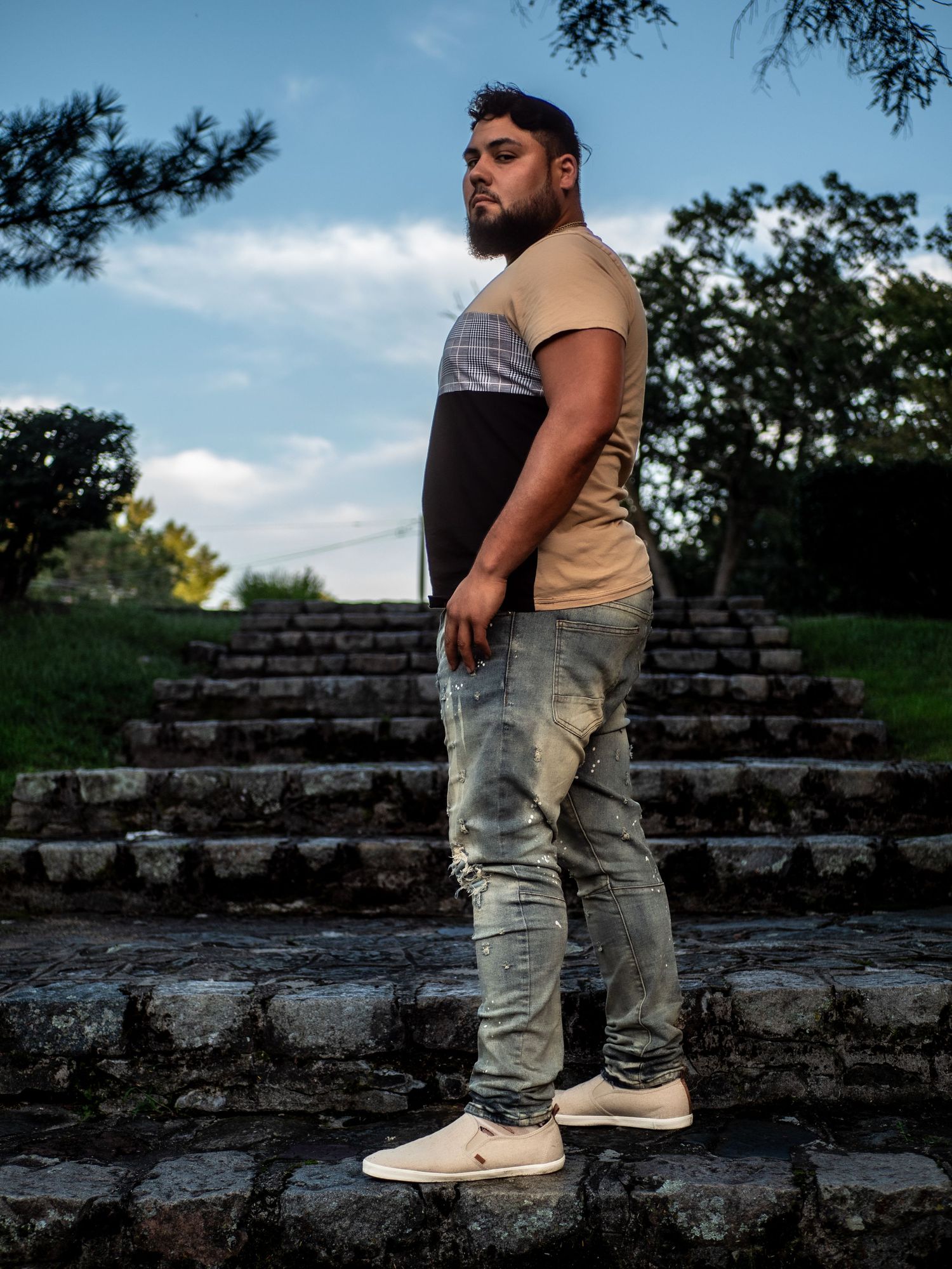 A person in a casual outfit stands on stone steps outdoors with trees and blue sky in the background.