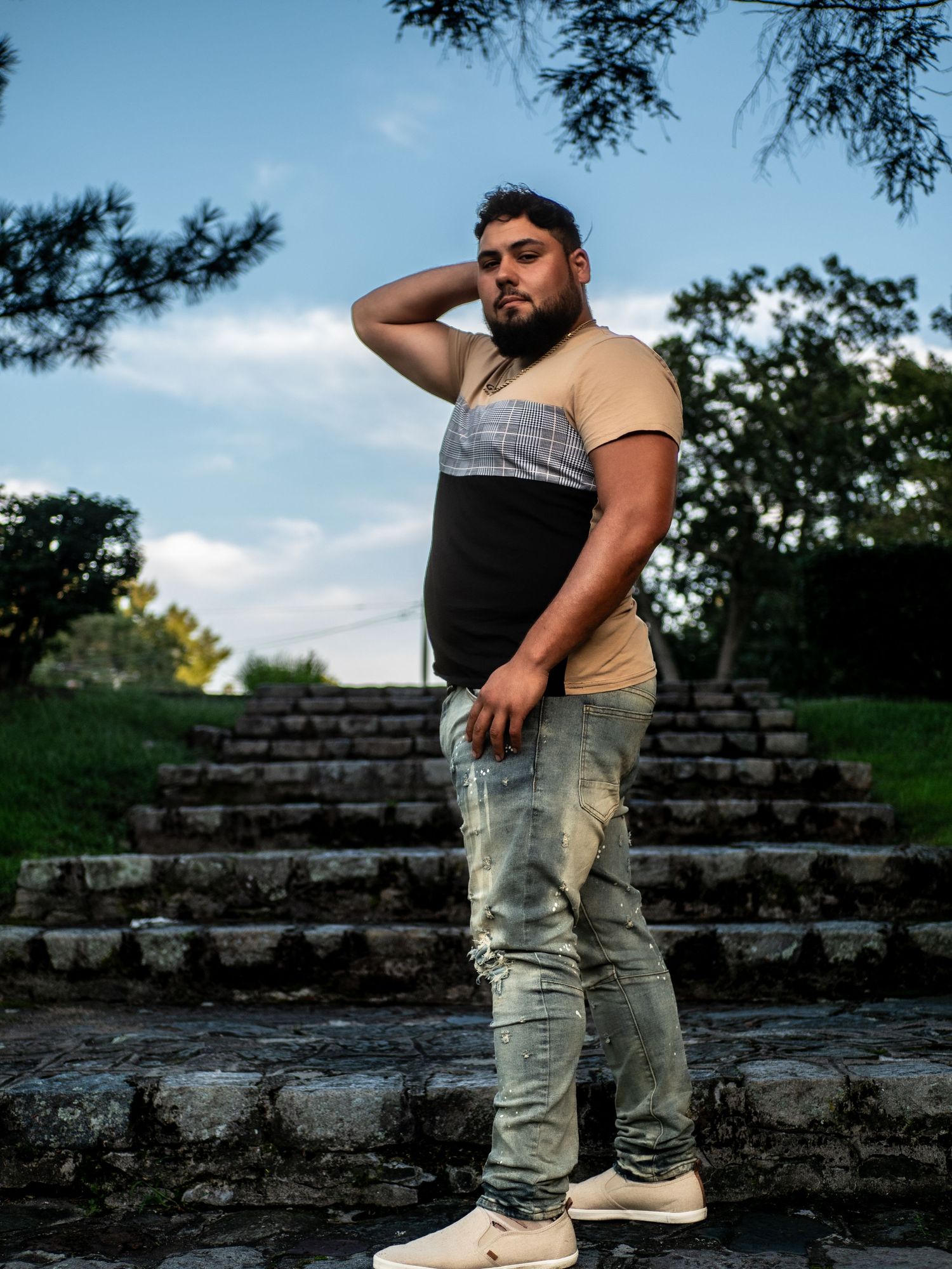 Someone poses on stone steps outdoors wearing a tan t-shirt and light jeans against a blue sky backdrop.