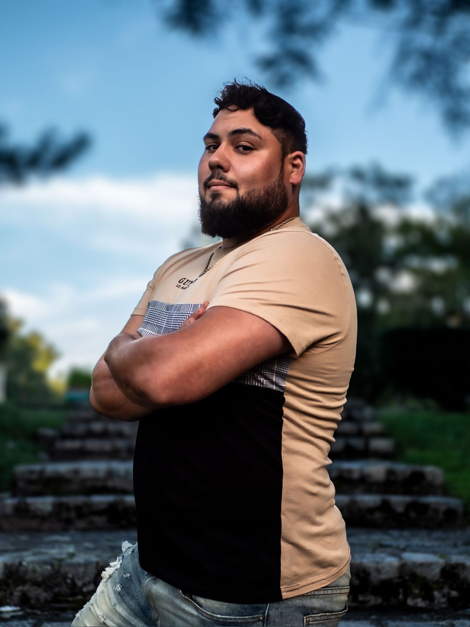 A person in a two-tone tan and black t-shirt poses confidently with crossed arms on stone steps during golden hour lighting.