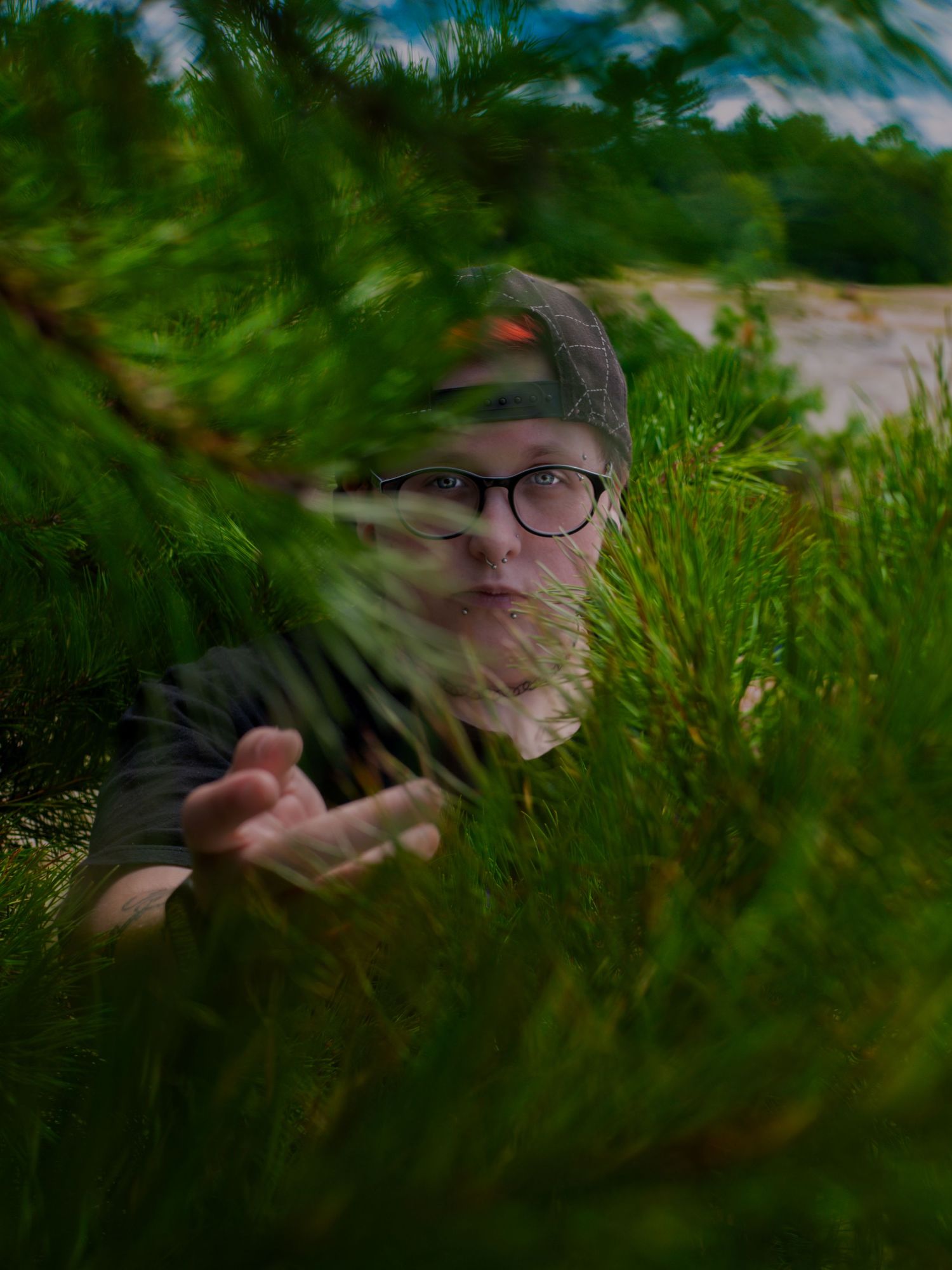 Someone wearing glasses and a gray cap peers through green foliage in an outdoor nature setting.