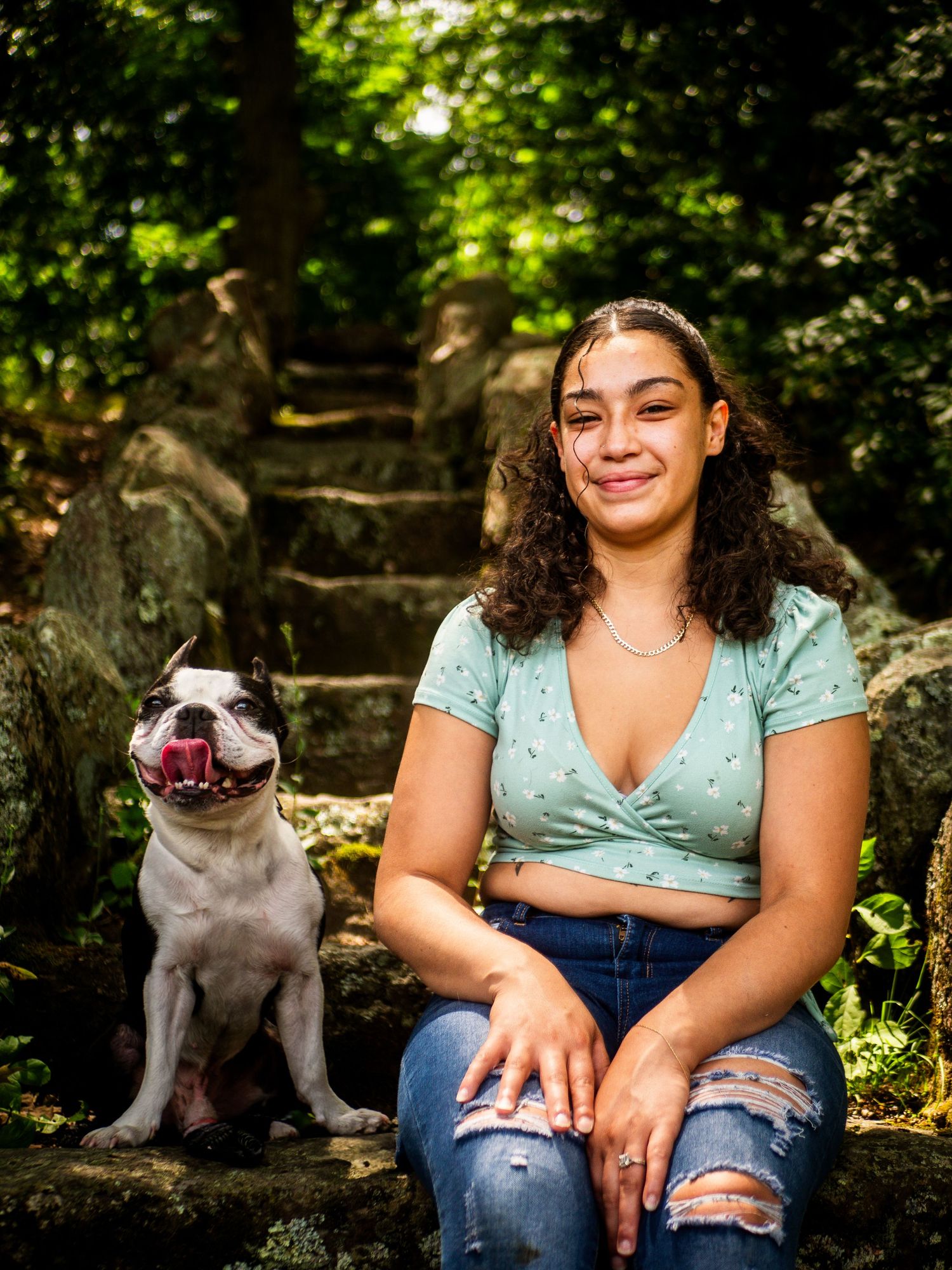 A happy person and their smiling pit bull sitting together on stone steps in a lush forest setting.