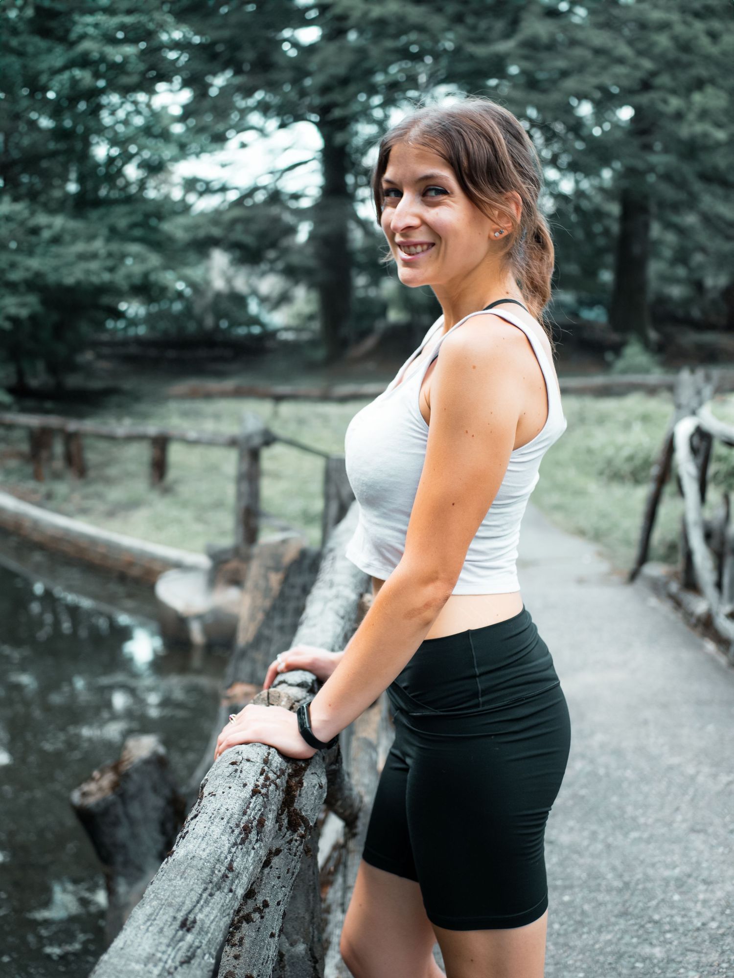 Someone wearing athletic attire smiles while standing on a wooded path next to a small stream or pond.