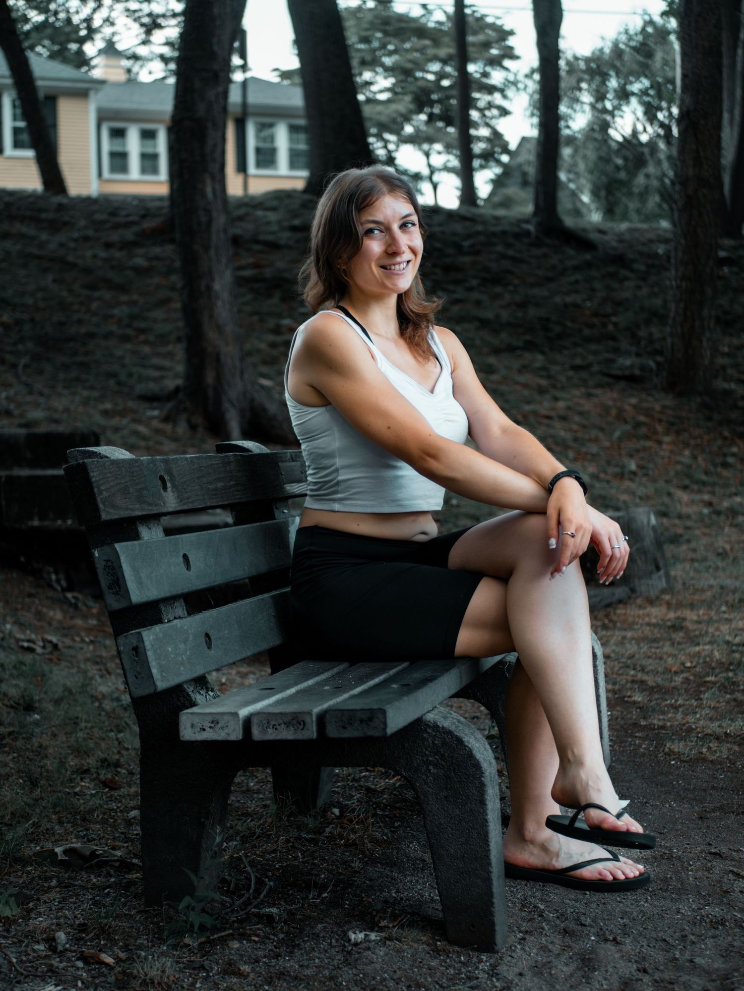 A person in a tank top and shorts sits relaxed on a wooden bench in a wooded outdoor setting at dusk.