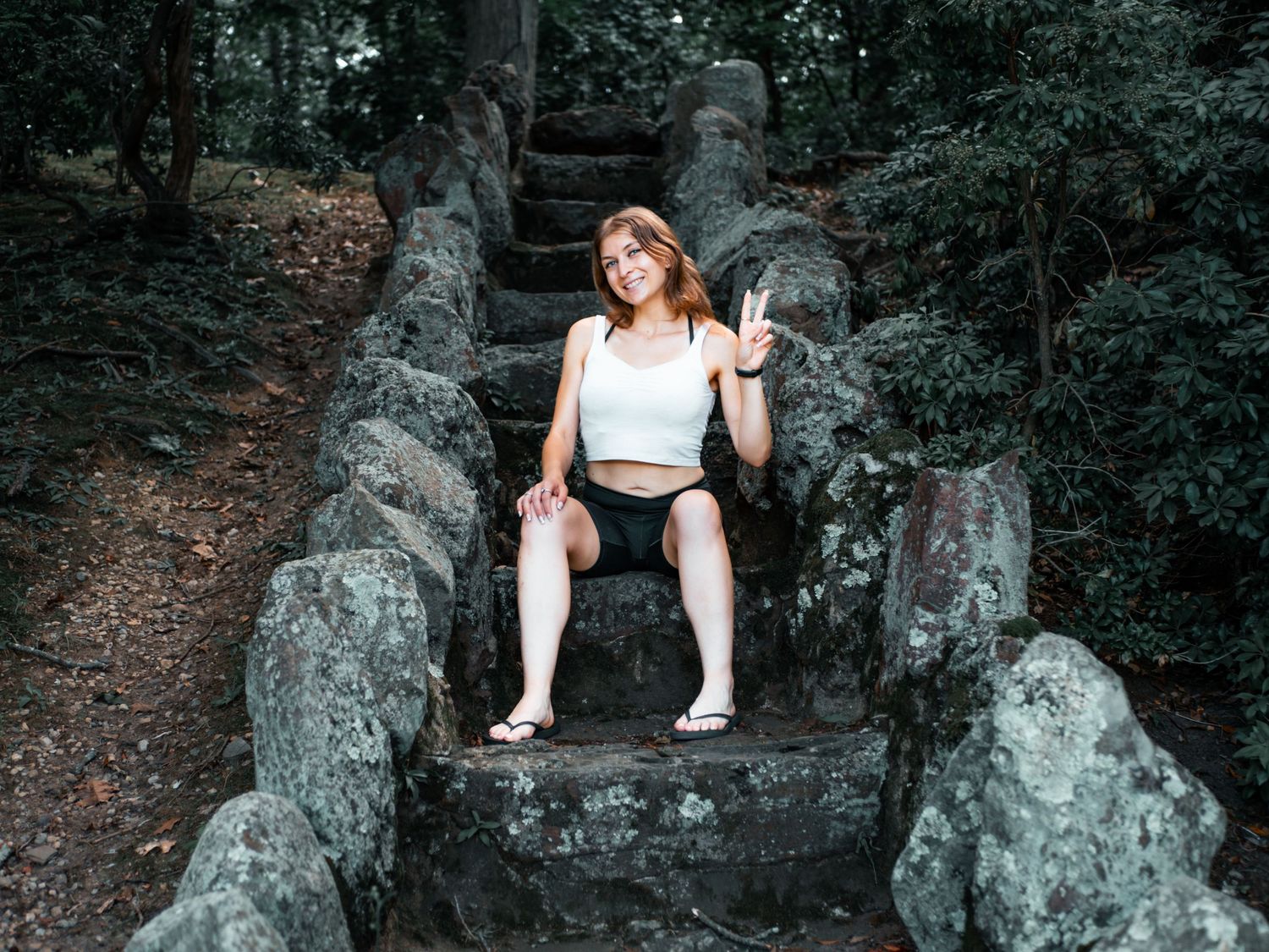 A person in a white crop top sits on ancient stone steps surrounded by lush forest vegetation.