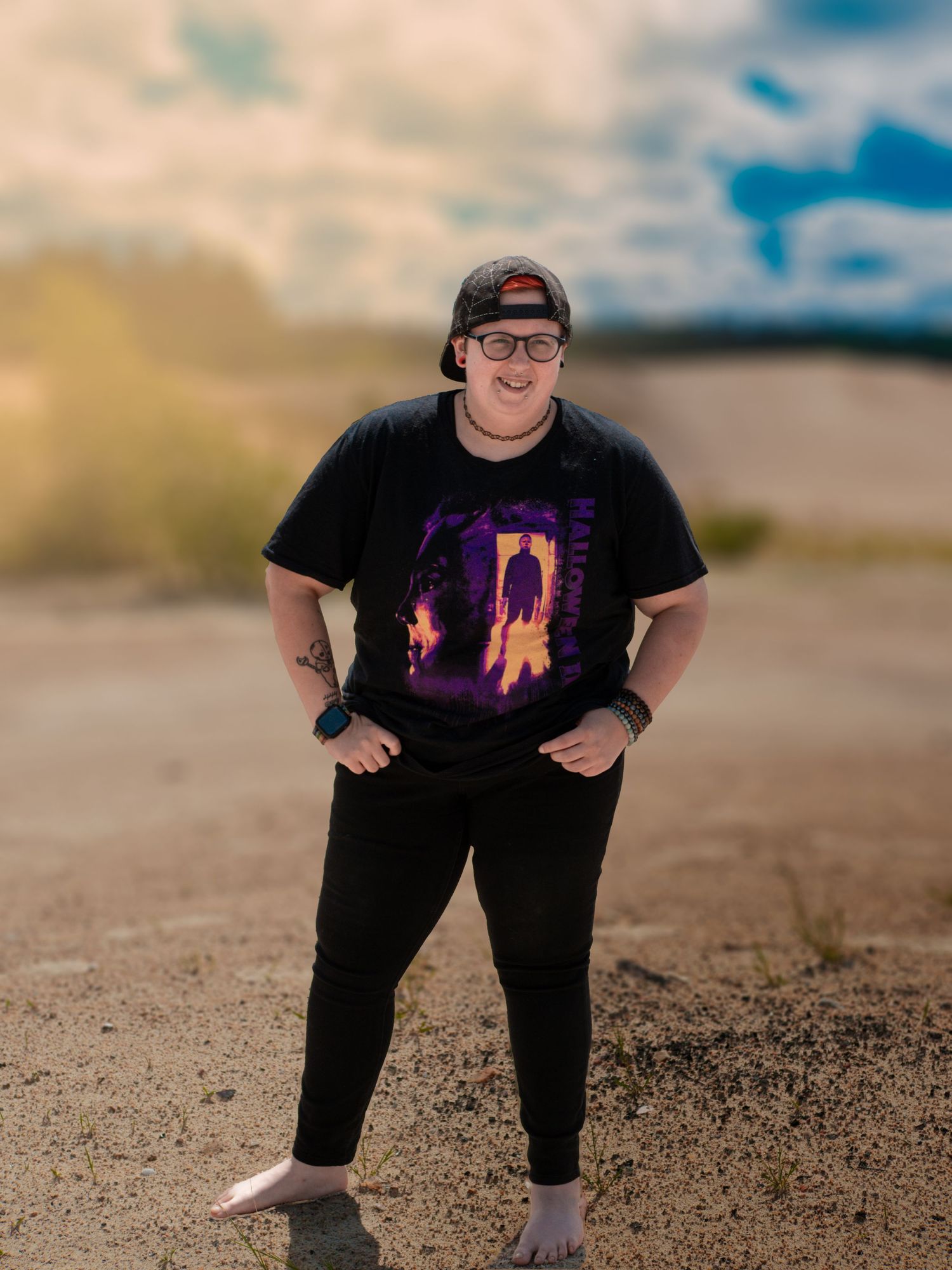 Someone wearing a black t-shirt with a colorful graphic design stands on sandy ground against a cloudy sky background.