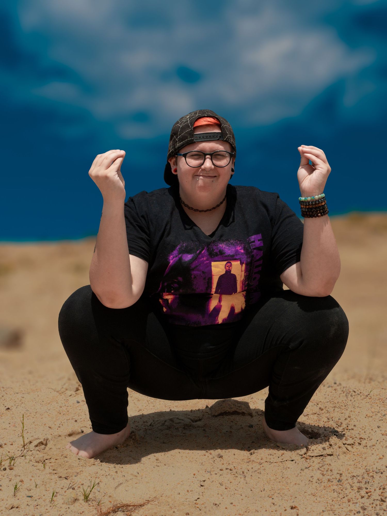 A person in a black t-shirt and cap squats on sand dunes under a blue cloudy sky.