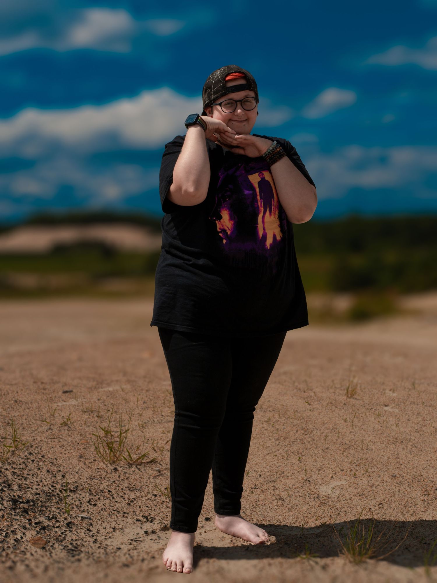 A person wearing all black stands barefoot on sand against a blue sky with white clouds.
