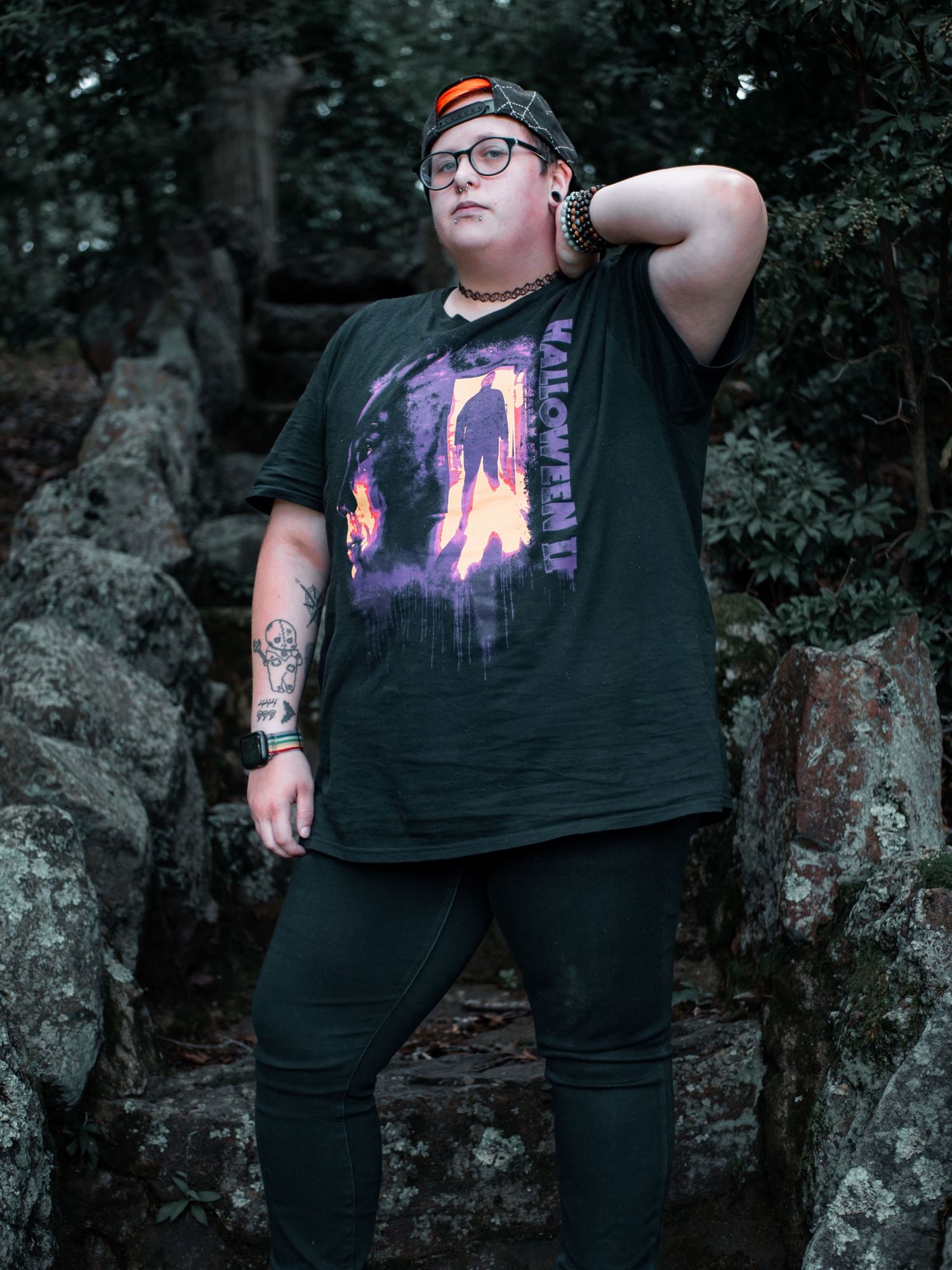 Person in black graphic t-shirt and pants poses against rock wall in outdoor setting with trees in background.