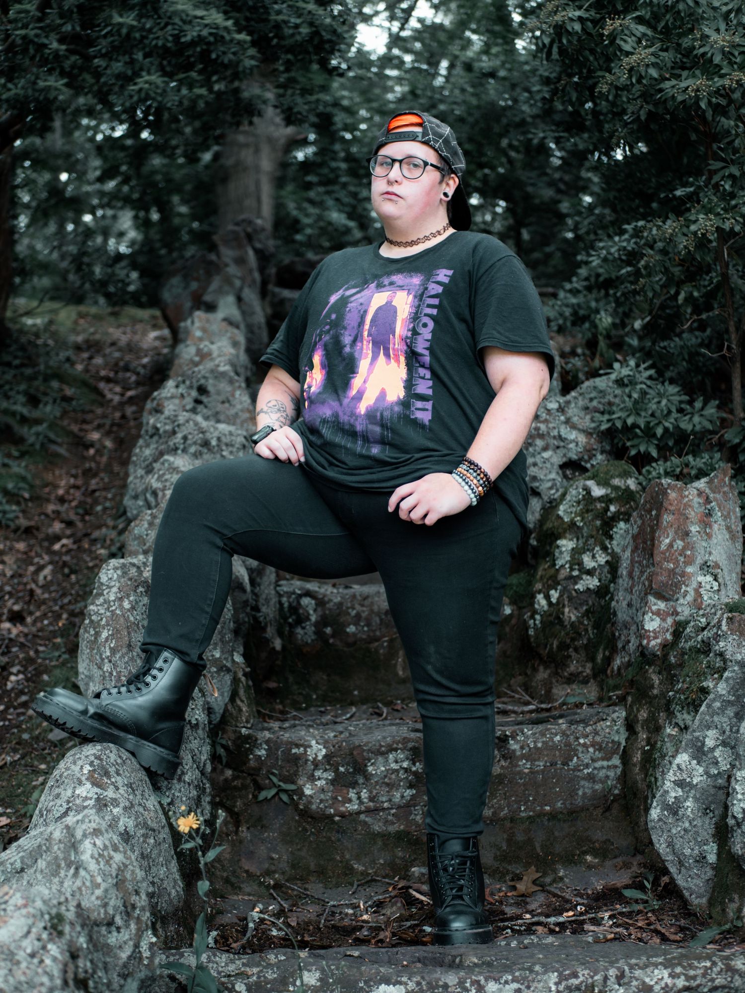 Someone wearing a graphic t-shirt, baseball cap and boots sits casually on large rocks in a wooded outdoor setting.