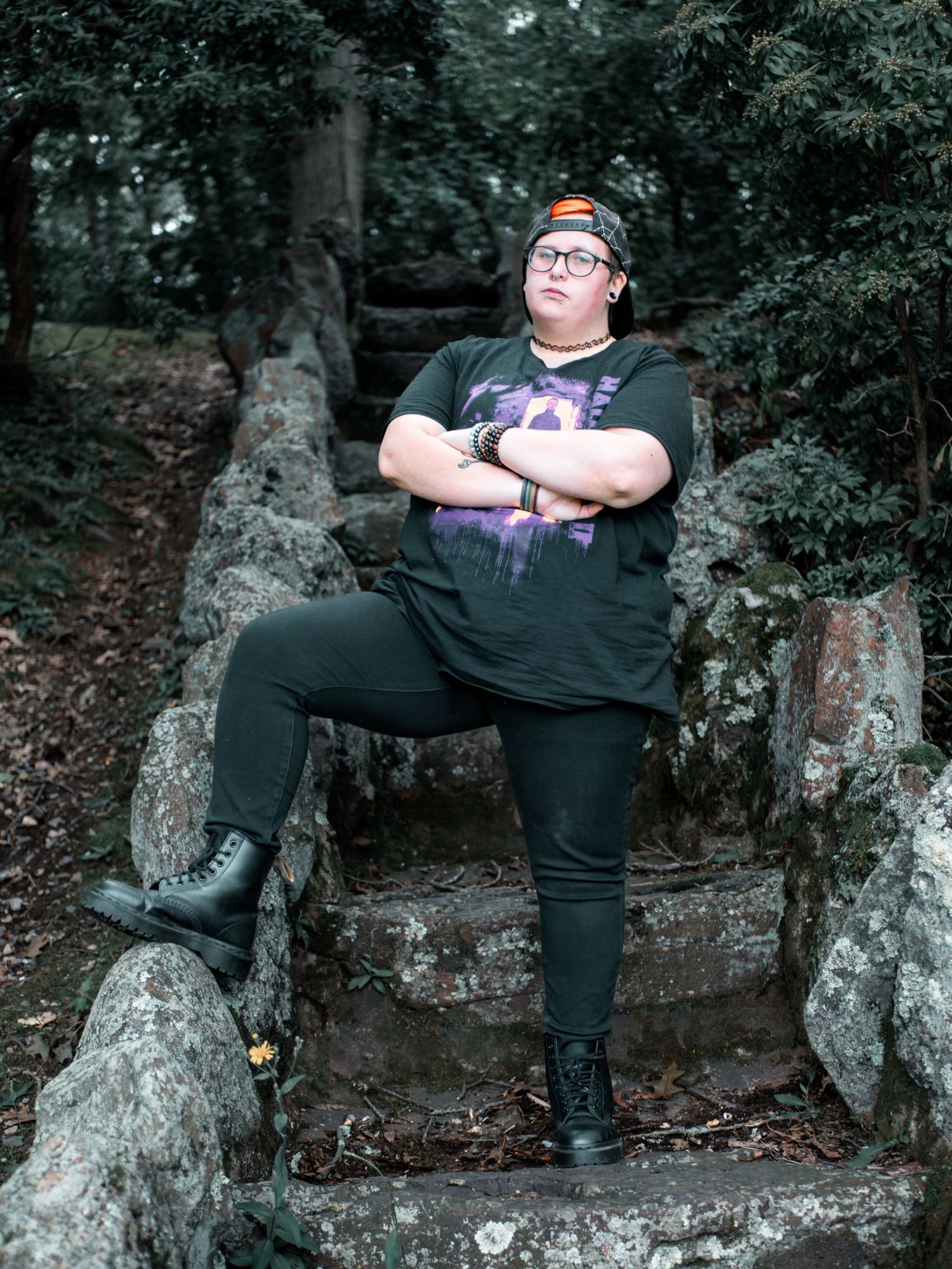 Person in black outfit and orange beanie sits confidently on stone steps in a wooded outdoor setting.