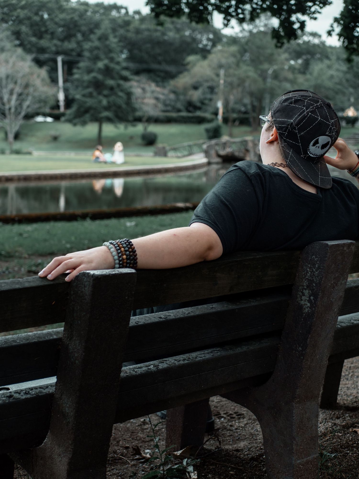 Person wearing a baseball cap sits alone on a wooden bench near a pond in a serene park setting.