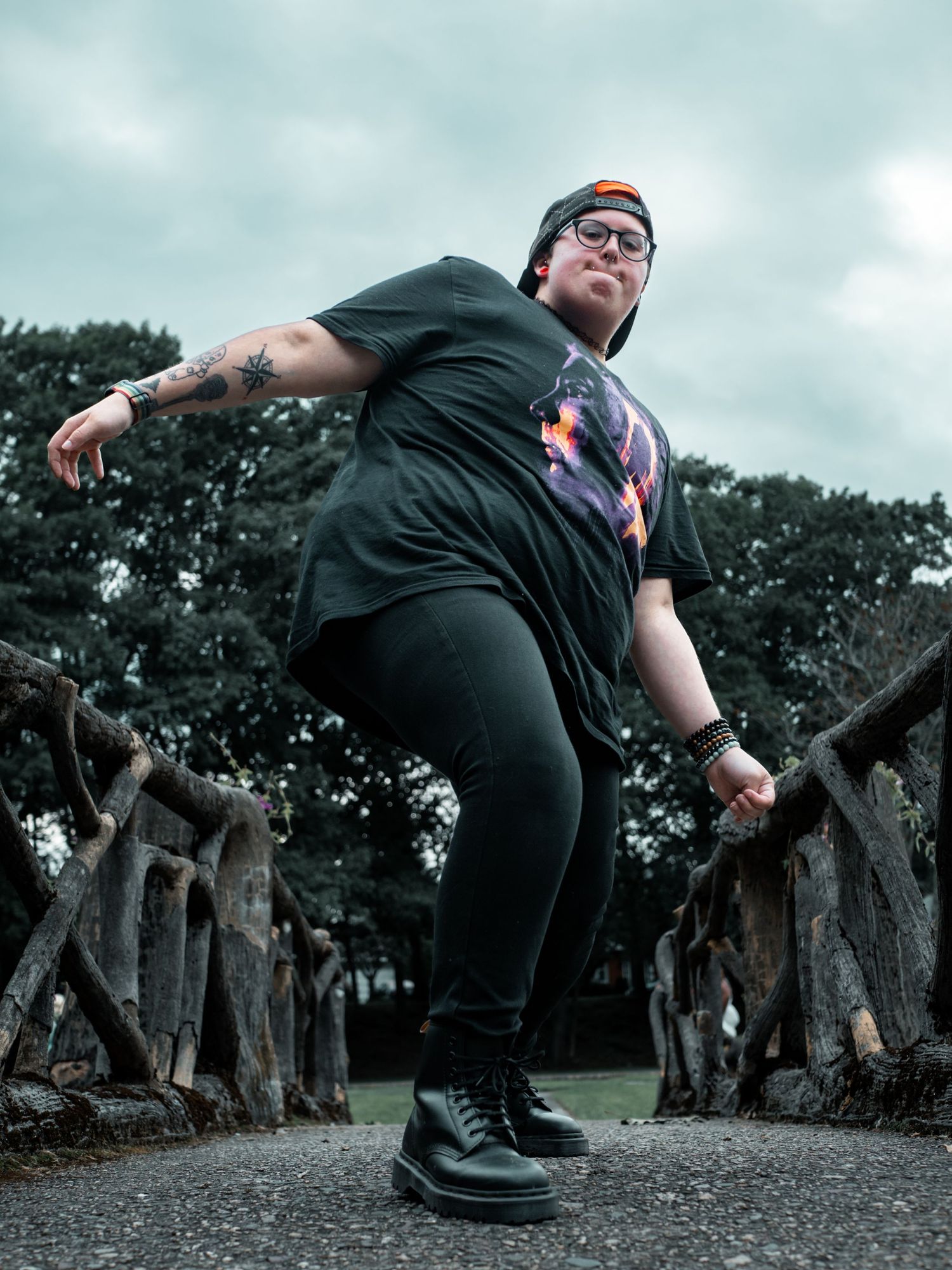 A person in dark clothing strikes an energetic dance pose on a wooden bridge in a dramatic outdoor setting.