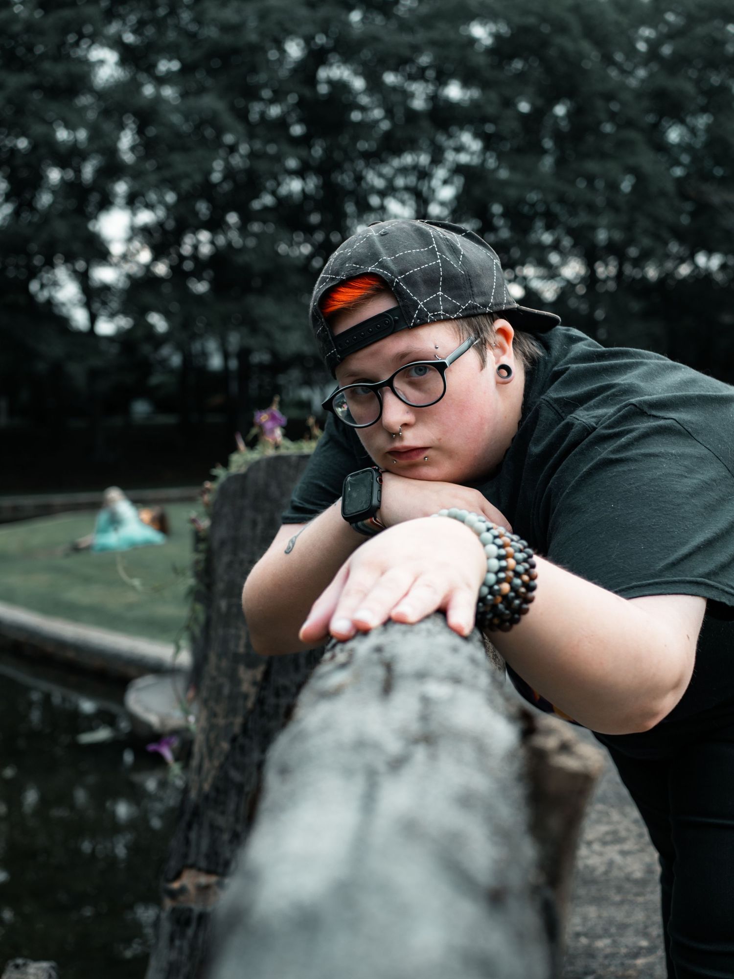 Someone wearing glasses and a baseball cap leans against a log railing in an outdoor setting with a moody atmosphere.