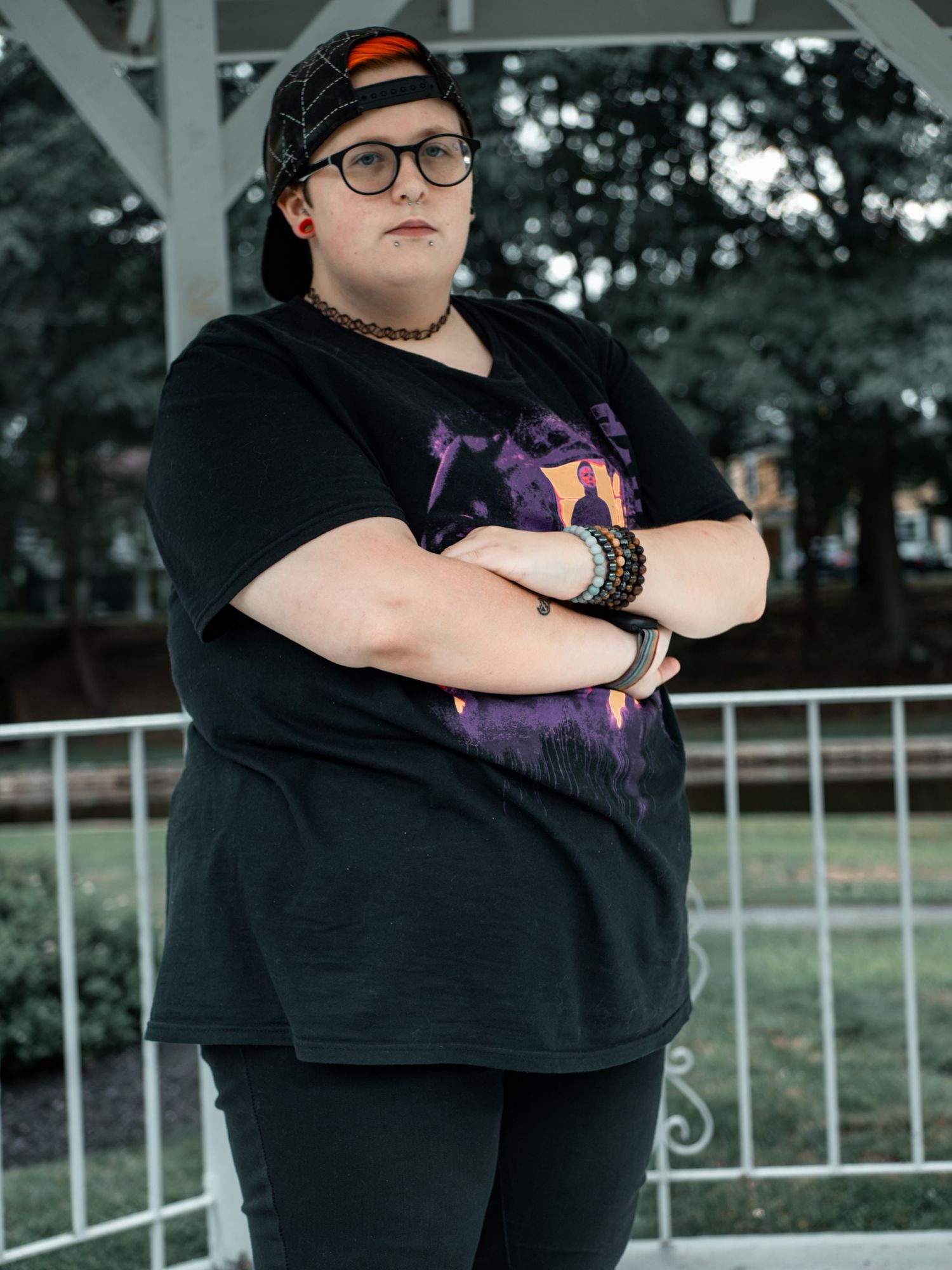 Individual wearing a black t-shirt, baseball cap and glasses poses with arms crossed against a railing outdoors.
