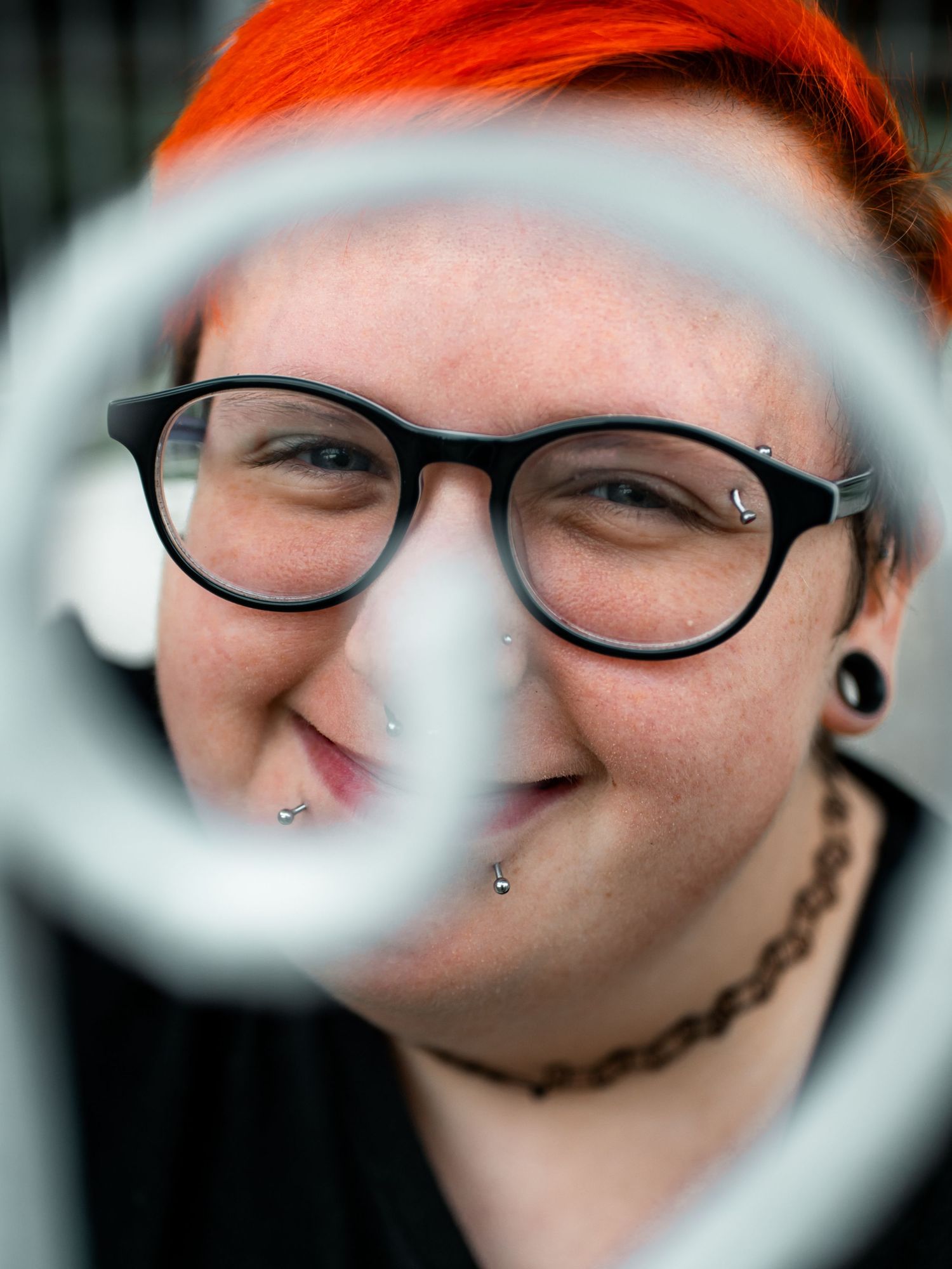 Close-up portrait shot through circular frame showing bright orange hair, black glasses and multiple facial piercings.