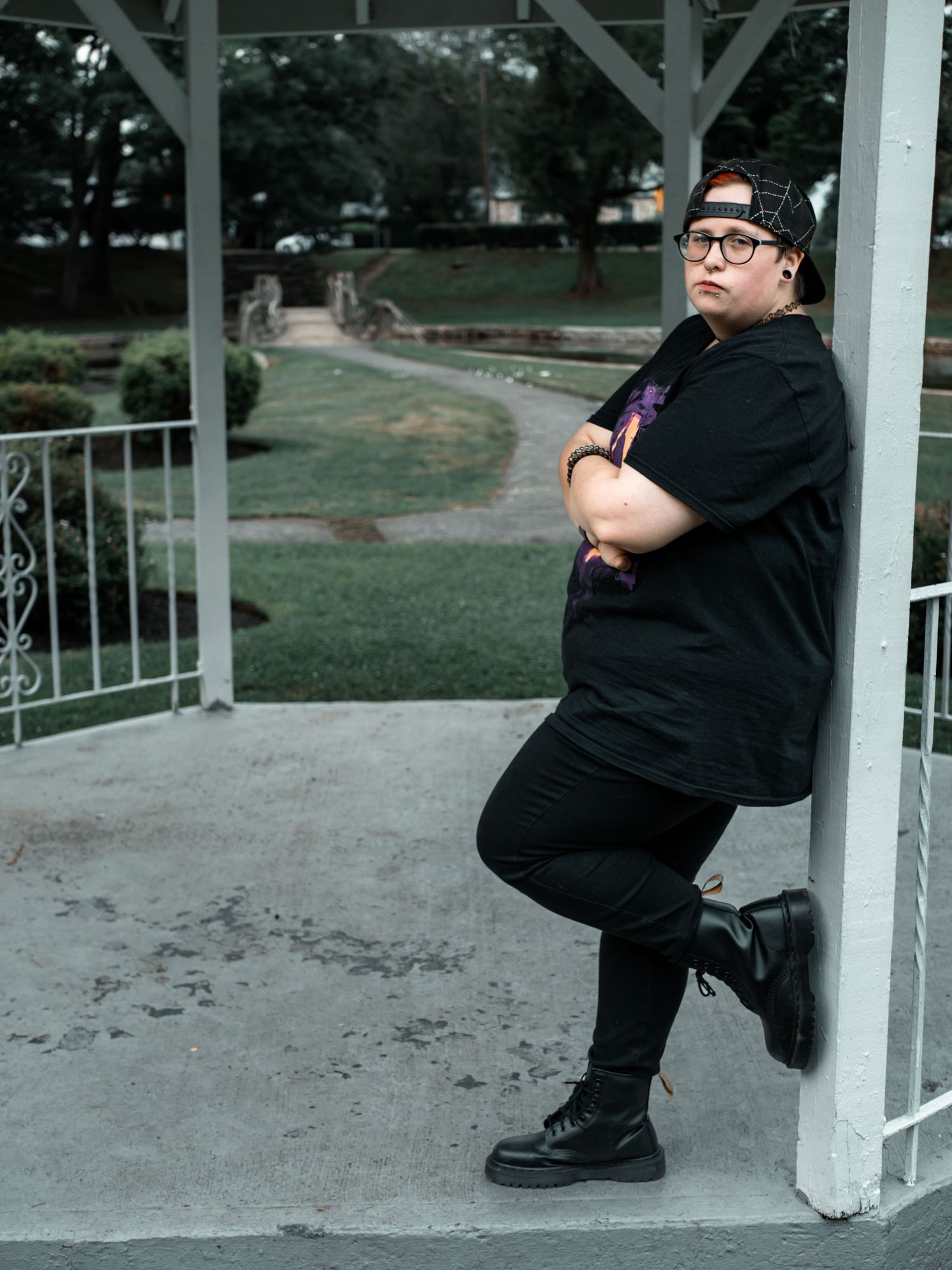 Person in all black attire poses casually leaning against white gazebo post in park setting.