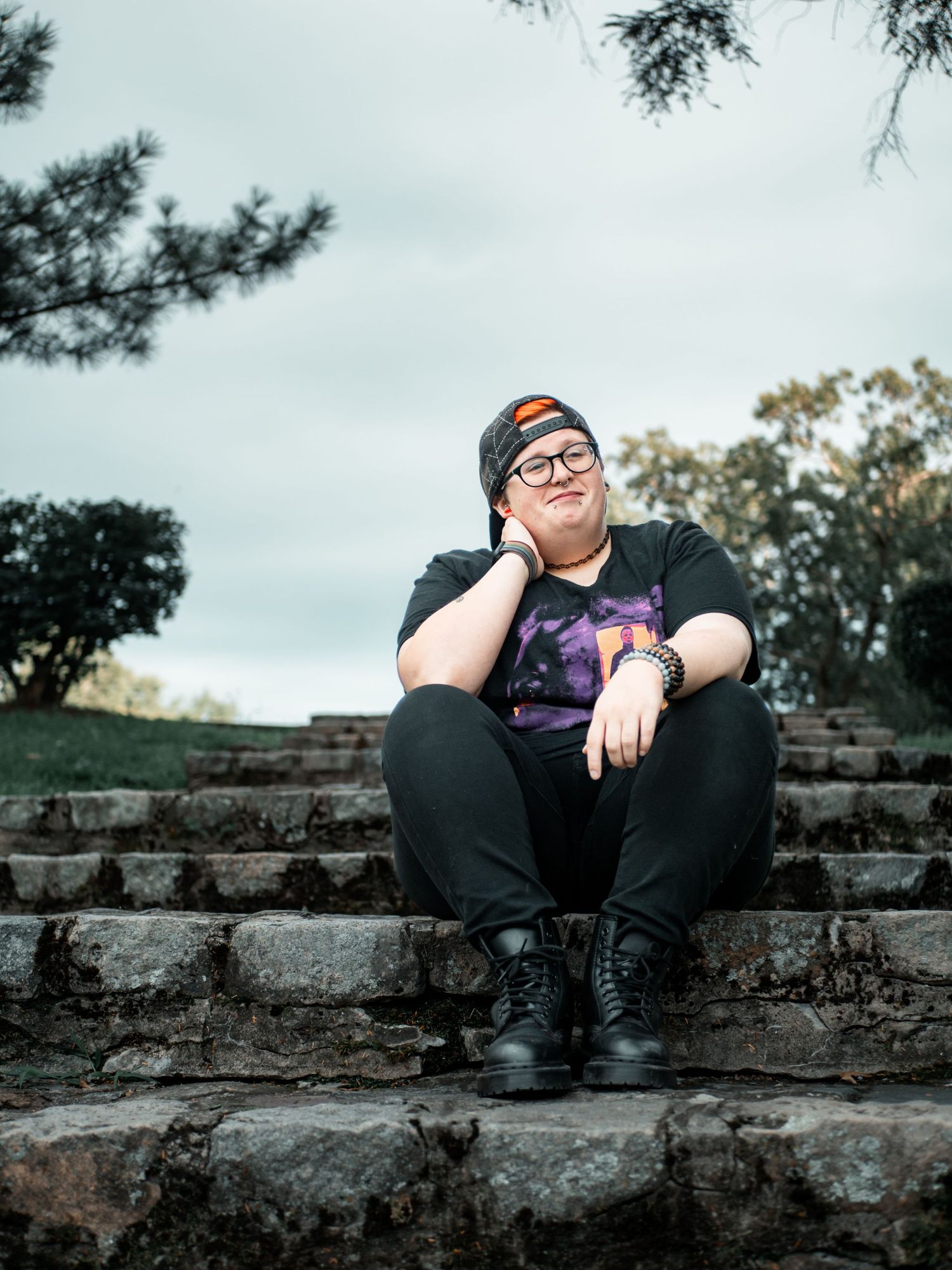 A person wearing glasses and a beanie sits on stone steps outdoors against a cloudy sky background.