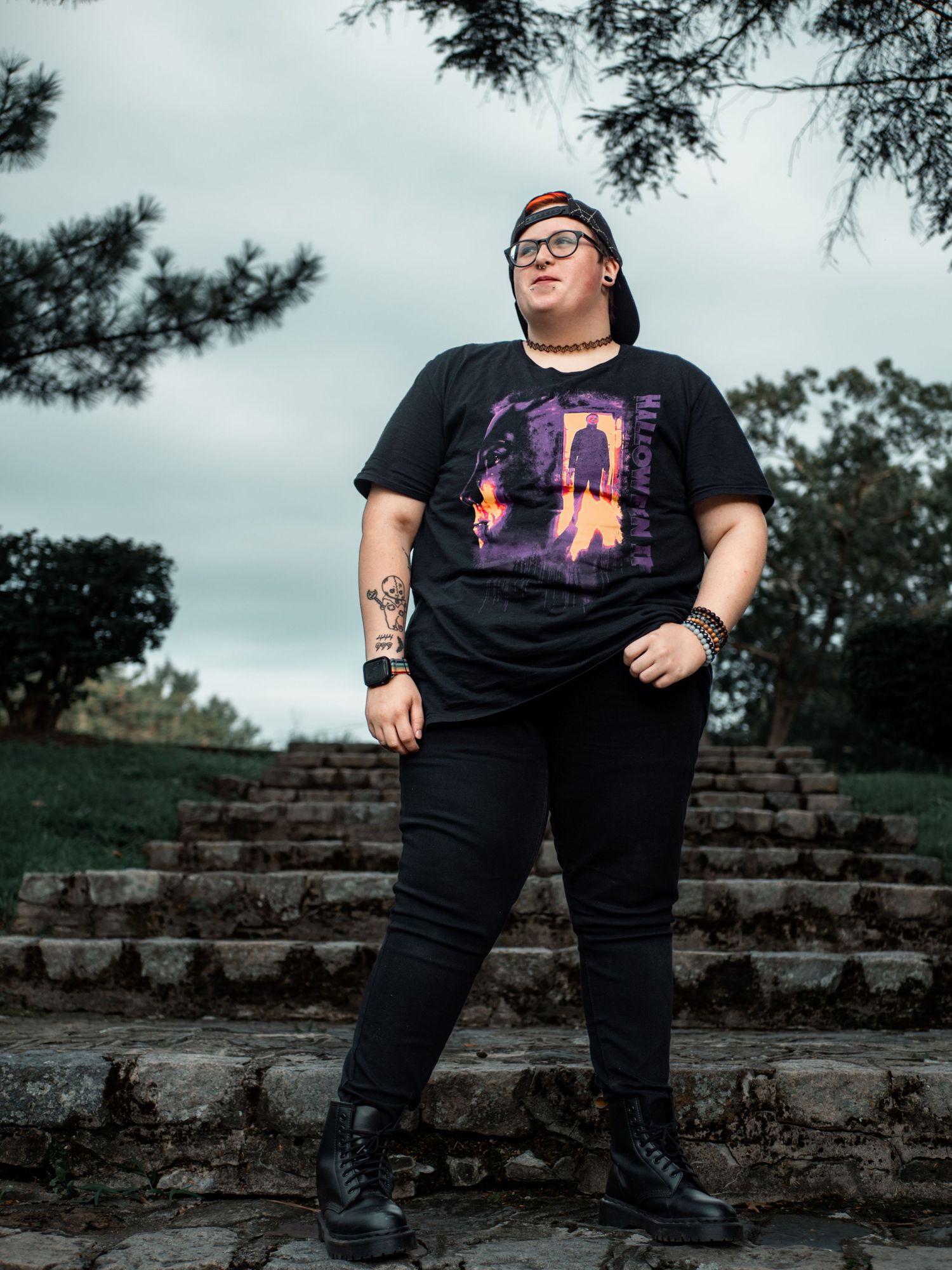 A person in black clothing and glasses poses on stone steps against a cloudy sky with pine trees in the background.