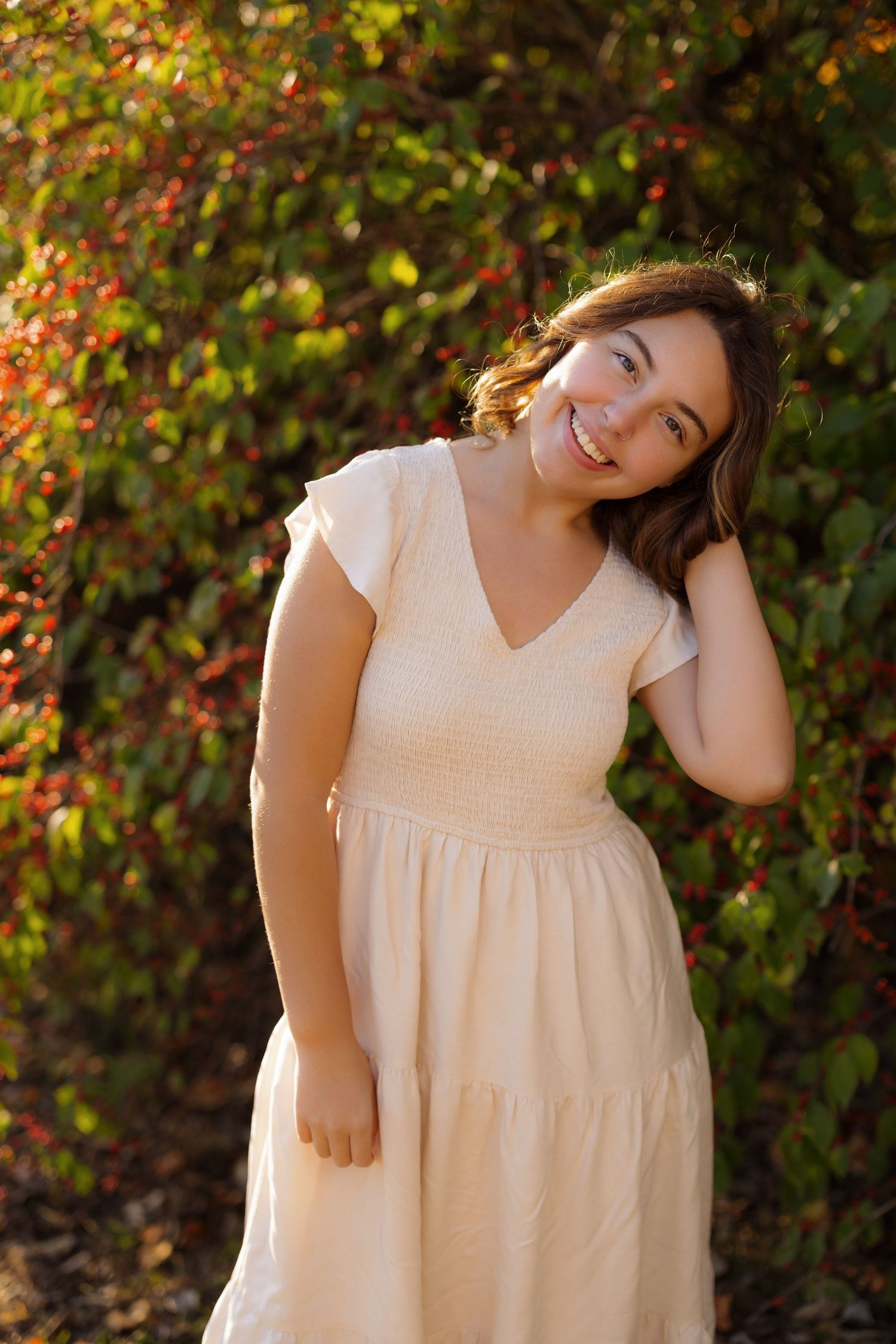 Portrait of someone in a cream-colored tiered dress posing outdoors against autumn foliage with warm sunlight.