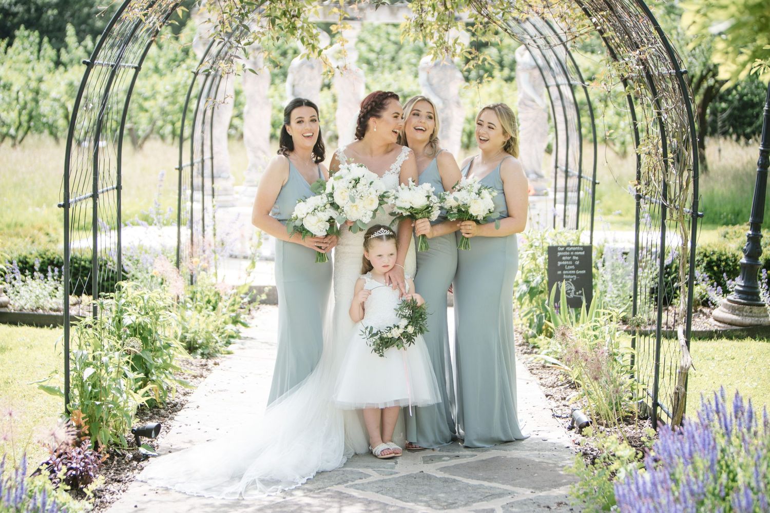 A bridal party in light green dresses poses under an arched garden trellis surrounded by blooming lavender at Tangled Garden wedding venue.
