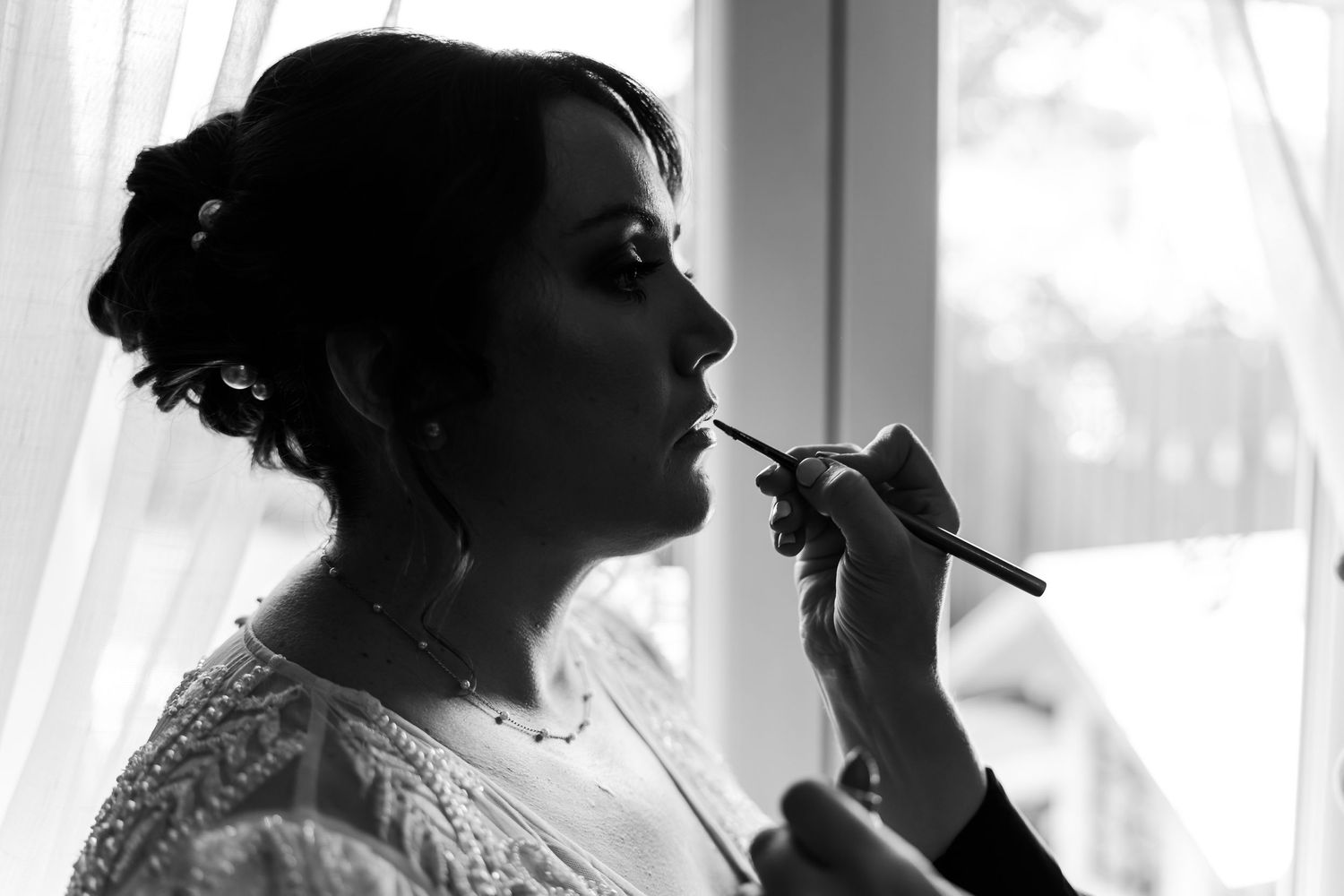 A silhouetted profile in black and white shows someone getting their makeup done near a window with soft light shining through.