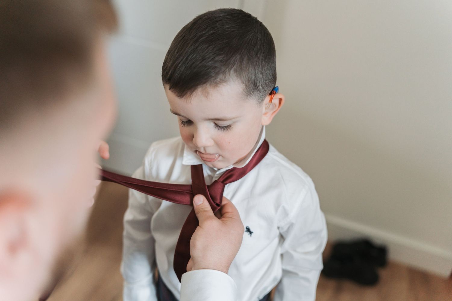 Someone helps adjusting a brown tie on a child wearing a crisp white dress shirt during formal preparation.