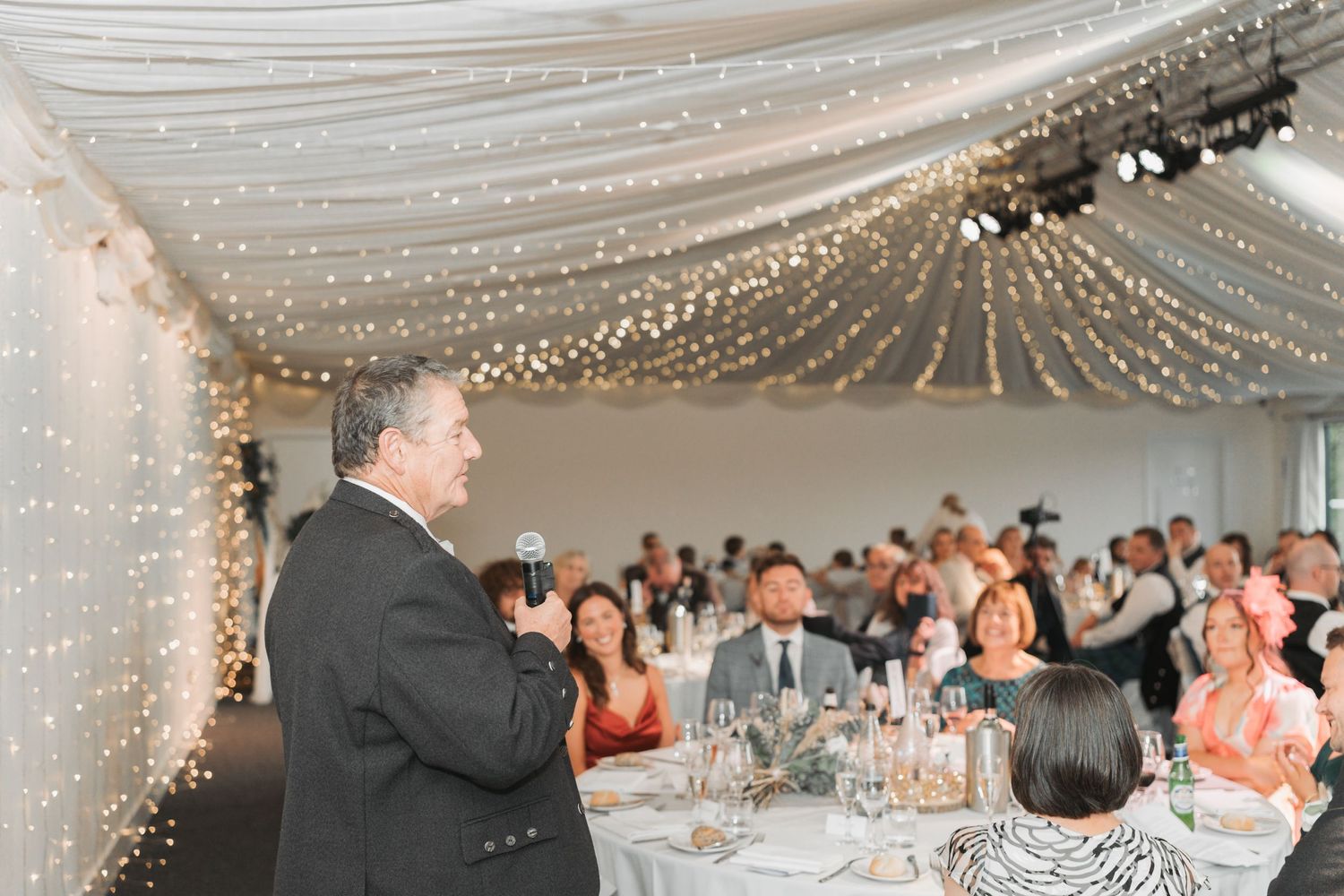 Wedding guests gather under a ceiling draped with twinkling lights as someone delivers a speech during the reception.