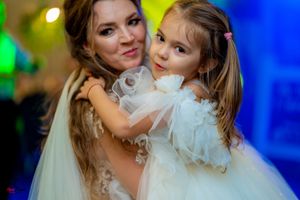 A bride in white lace dress embraces a young girl in a fluffy white dress at a wedding reception.