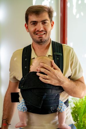 Person in overalls smiling and holding something while standing indoors.