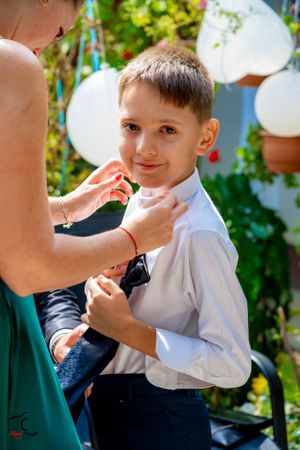 Person adjusting formal wear on child in white shirt and blue vest in garden setting.