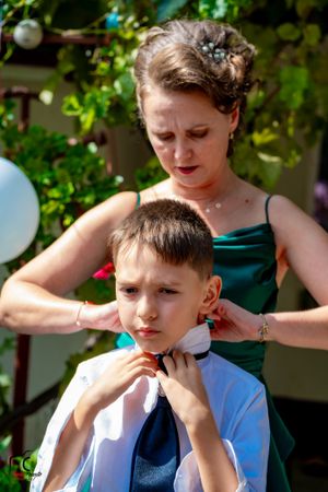 Person in white shirt and blue vest being helped with formal attire outdoors.