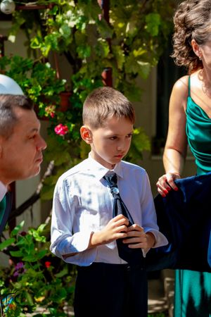 Child in formal attire holding something while standing in garden setting with green foliage.