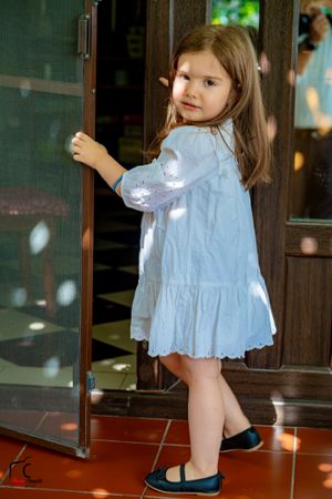 Child in white dress playing near wooden screen door with sunlight casting shadows.