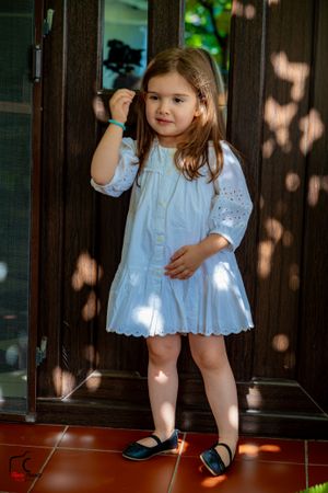 Child wearing white eyelet dress and dark shoes standing in doorway with filtered sunlight.