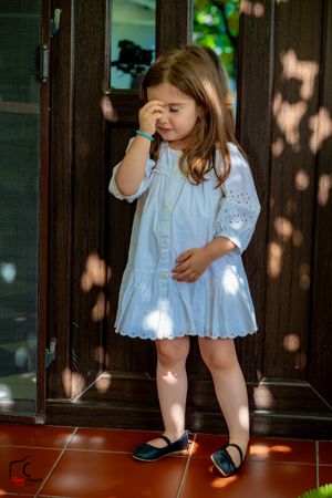 Little one in white cotton dress exploring shadowy porch area.